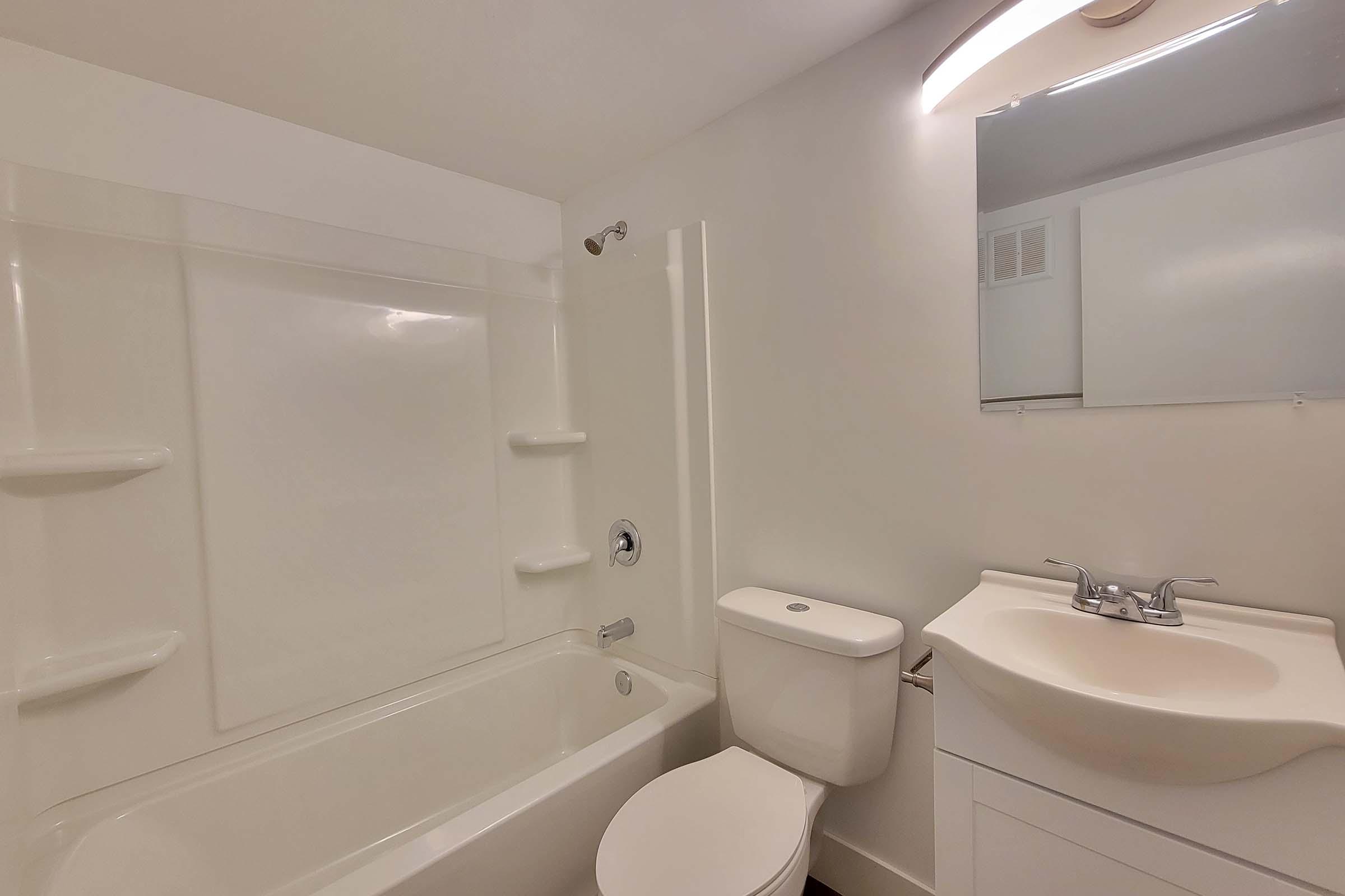 A modern bathroom featuring a bathtub with a showerhead, white walls, and a minimalist design. There is a toilet next to a sleek vanity with a sink and a mirror above. The overall color scheme is light and neutral, creating a clean and spacious appearance.