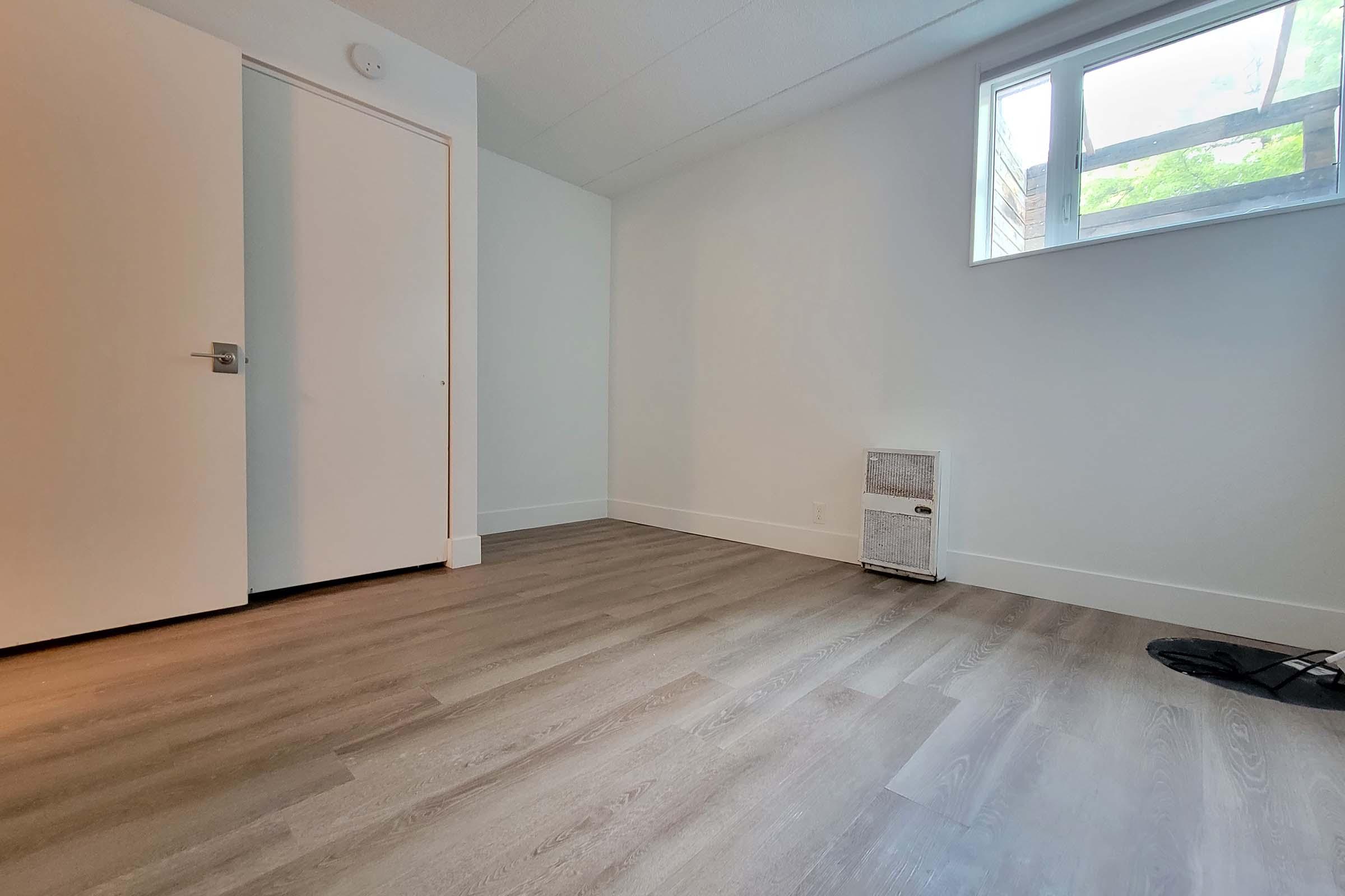 A minimalist room featuring light-colored walls, a window allowing natural light, a closed white door, and light wooden flooring. A small item resembling a heater or storage unit is positioned near the wall, and a black mat is located on the floor. The overall ambiance is bright and airy.