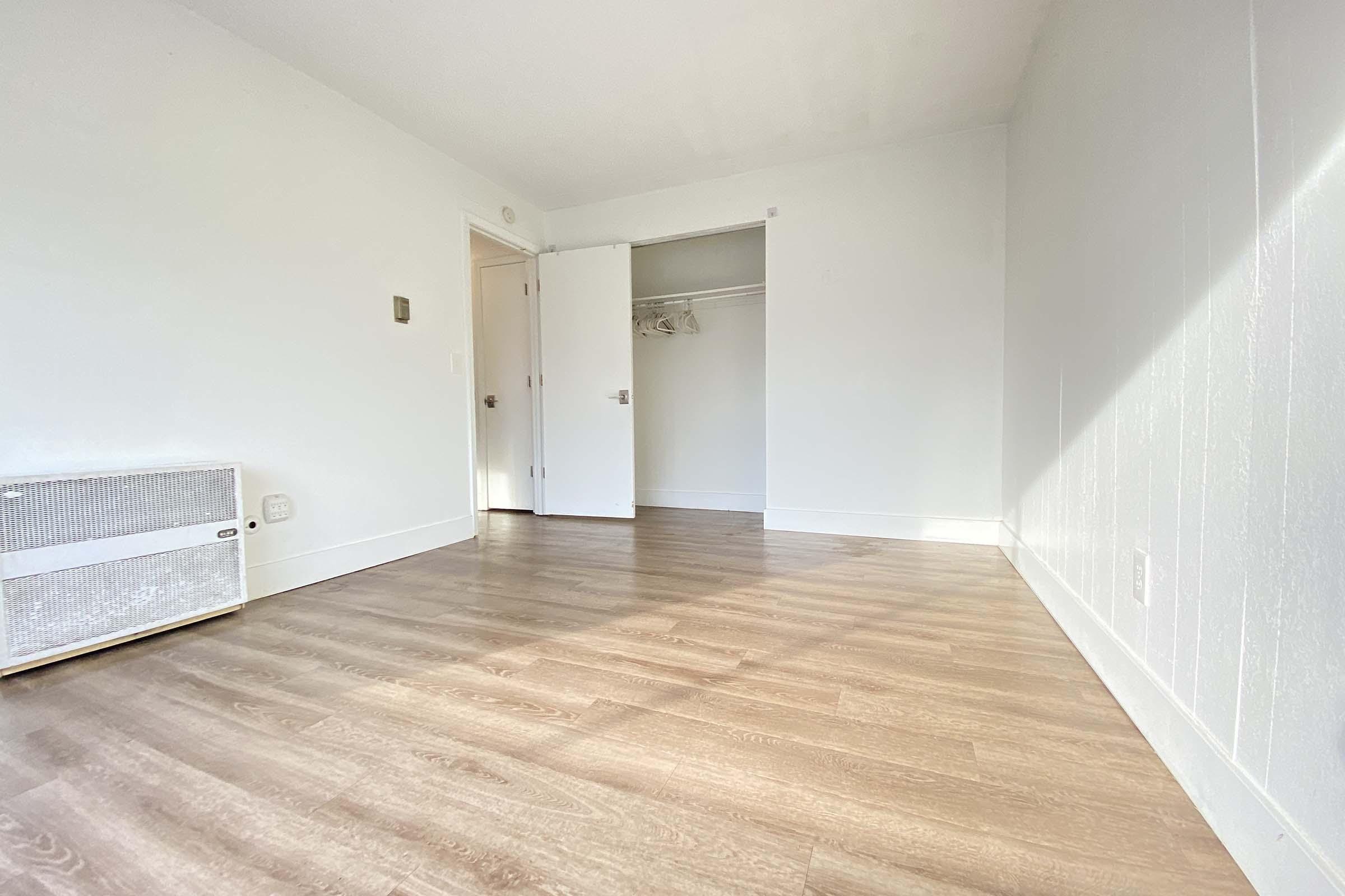 A spacious, bright, empty room with light-colored walls and wooden flooring. One corner features a wall heater. There's a closet with open doors, and a large door leading to another area is also slightly ajar, allowing natural light to stream in.