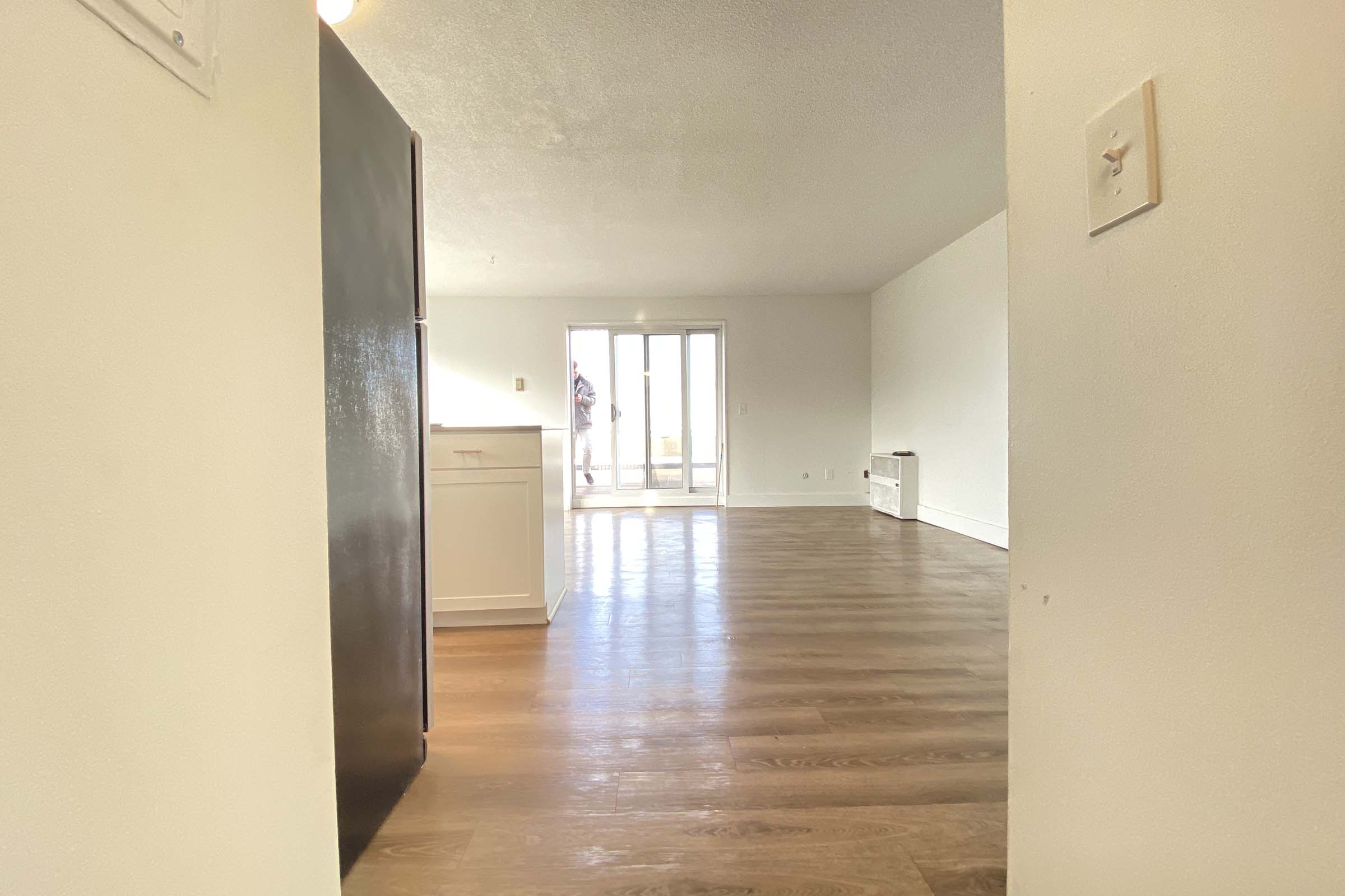 View of a spacious, well-lit room with hardwood floors, featuring a doorway leading to a balcony. The kitchen area is visible on the left, with a refrigerator and a doorway. Large windows allow natural light to fill the space, creating a welcoming atmosphere.