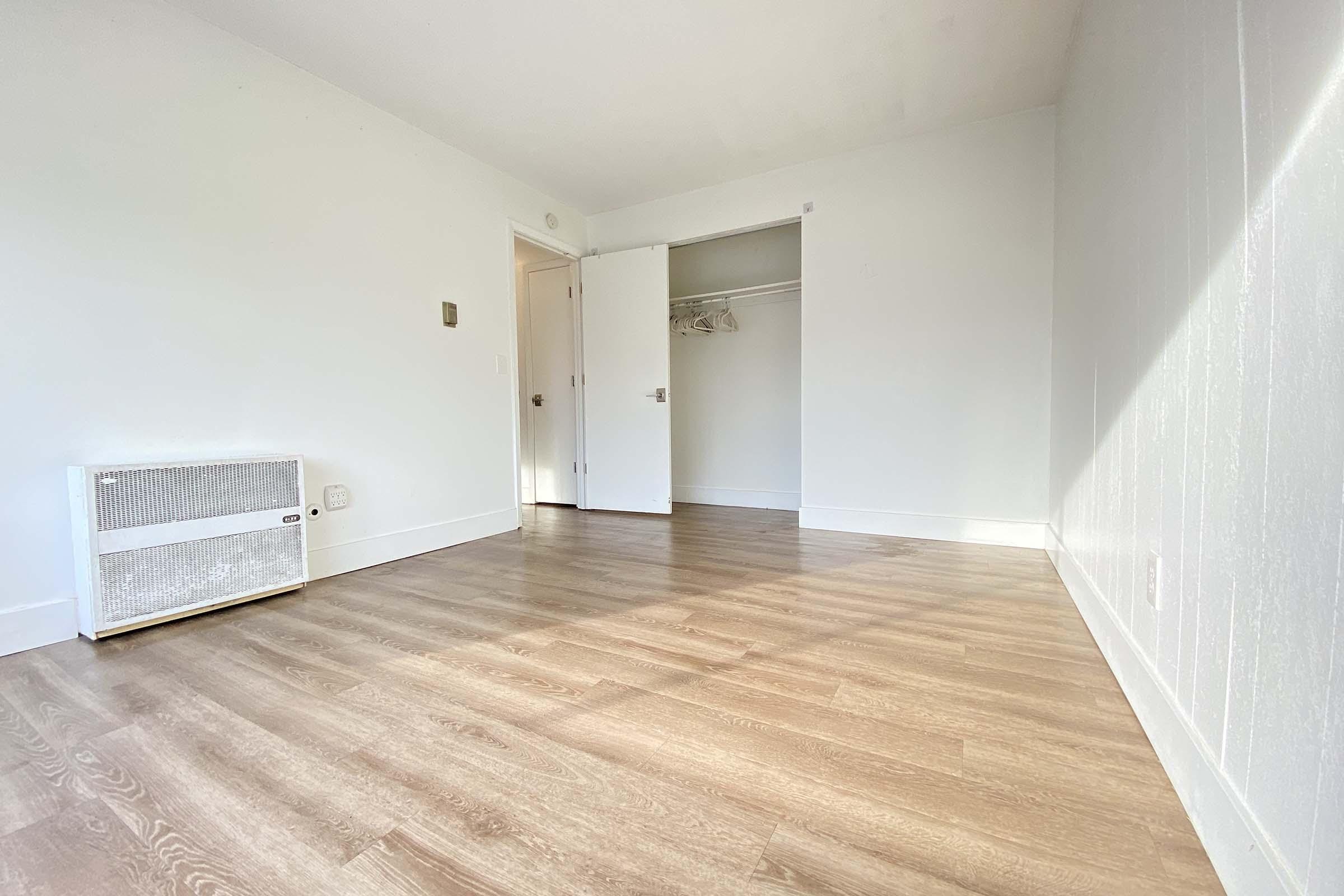 A bright, empty room with light-colored walls and hardwood flooring. There is a wall-mounted heater on the left, a partially open door leading to a closet, and ample sunlight illuminating the space from a window, creating a clean and spacious atmosphere.