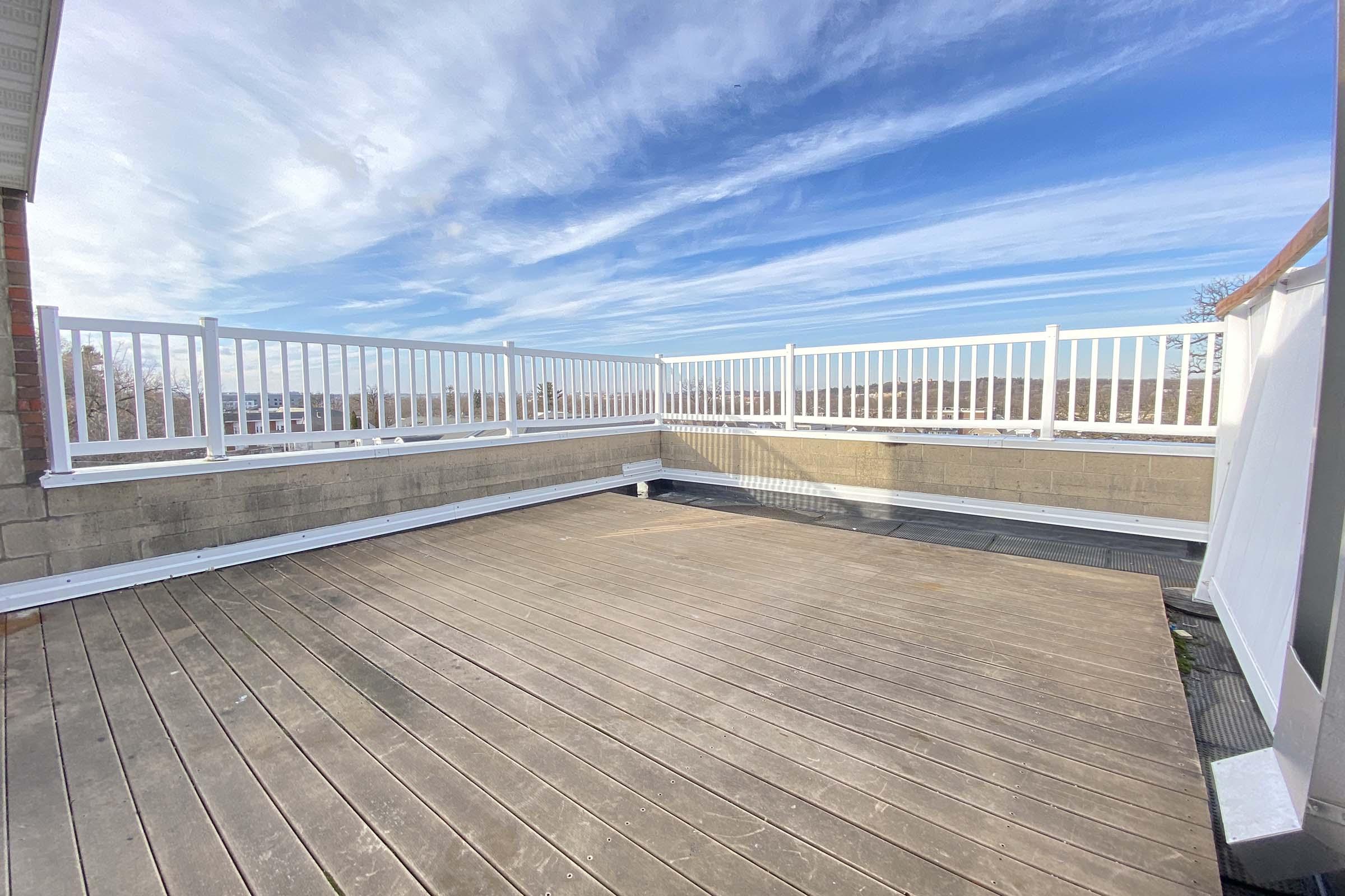 A spacious rooftop deck featuring a wooden floor and white railing, with a clear blue sky and wispy clouds in the background. The area appears clean and well-maintained, offering a panoramic view. The scene suggests a bright, sunny day, ideal for relaxation or gatherings.