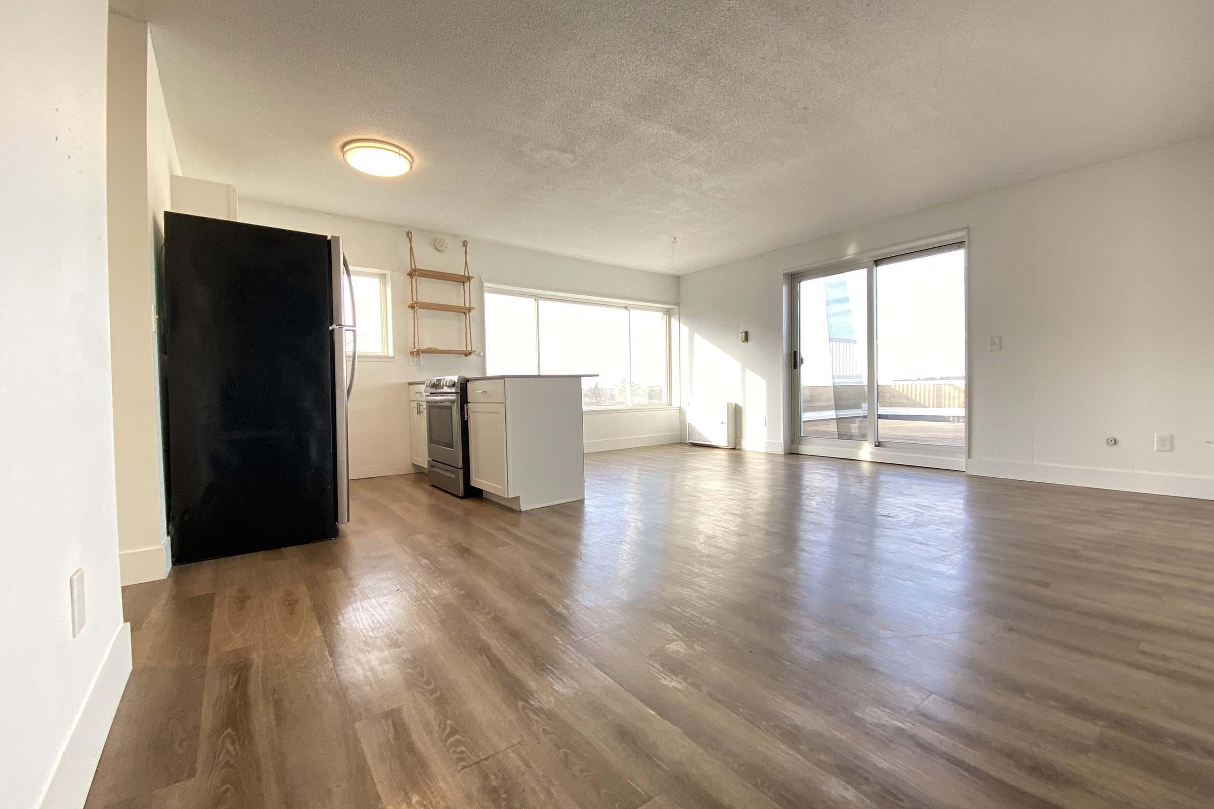 Spacious, bright interior of an apartment featuring a modern kitchen with a refrigerator and stove. Open layout with large windows allowing natural light, hardwood flooring throughout, and a sliding glass door leading to a balcony or outdoor space. Minimalist design with neutral colors.