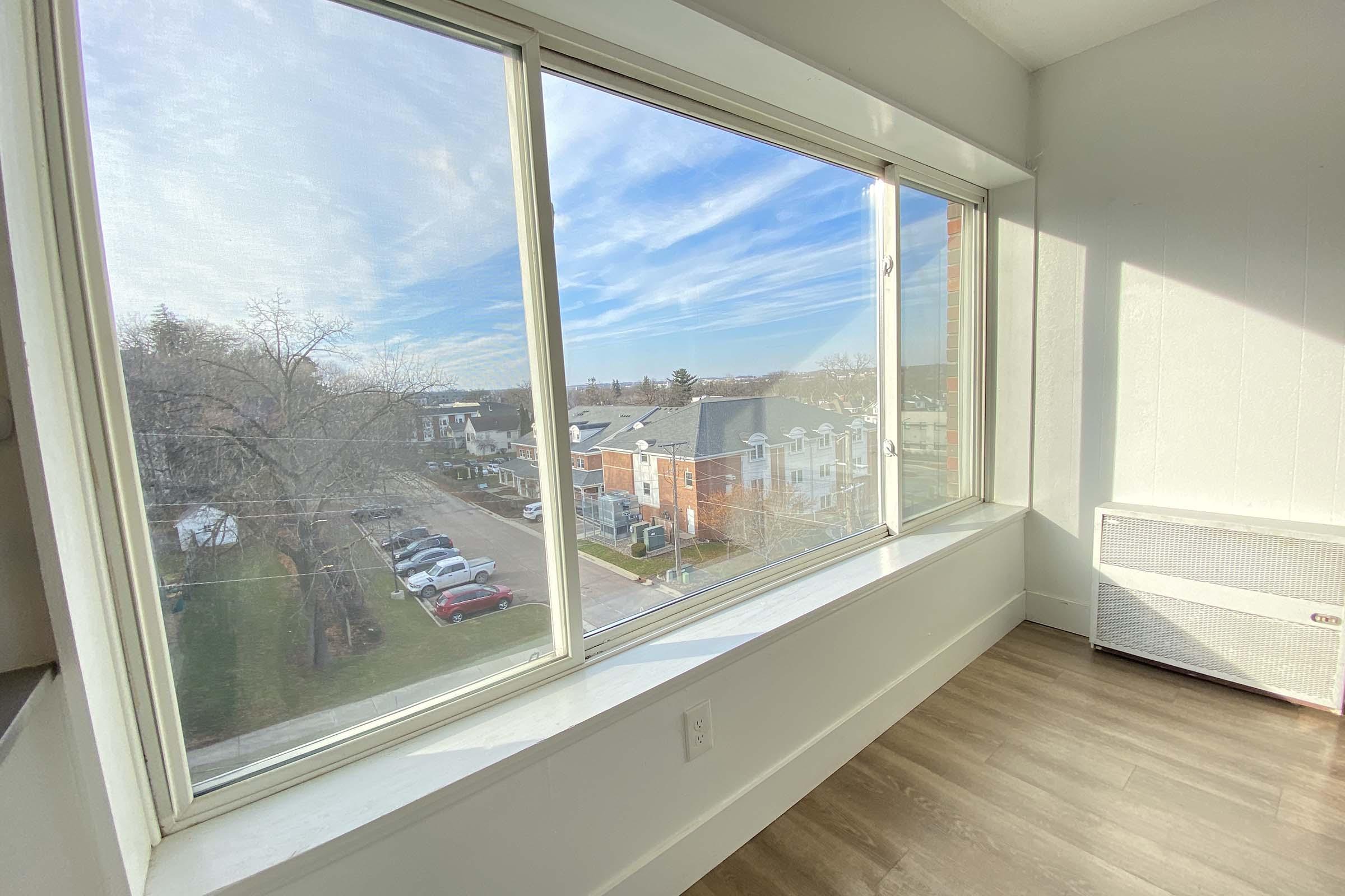 A bright room with large windows offering a view of a residential area. The scene includes parked cars along a lined street, trees, and nearby buildings under a partly cloudy sky. The interior features light-colored walls and wooden flooring.