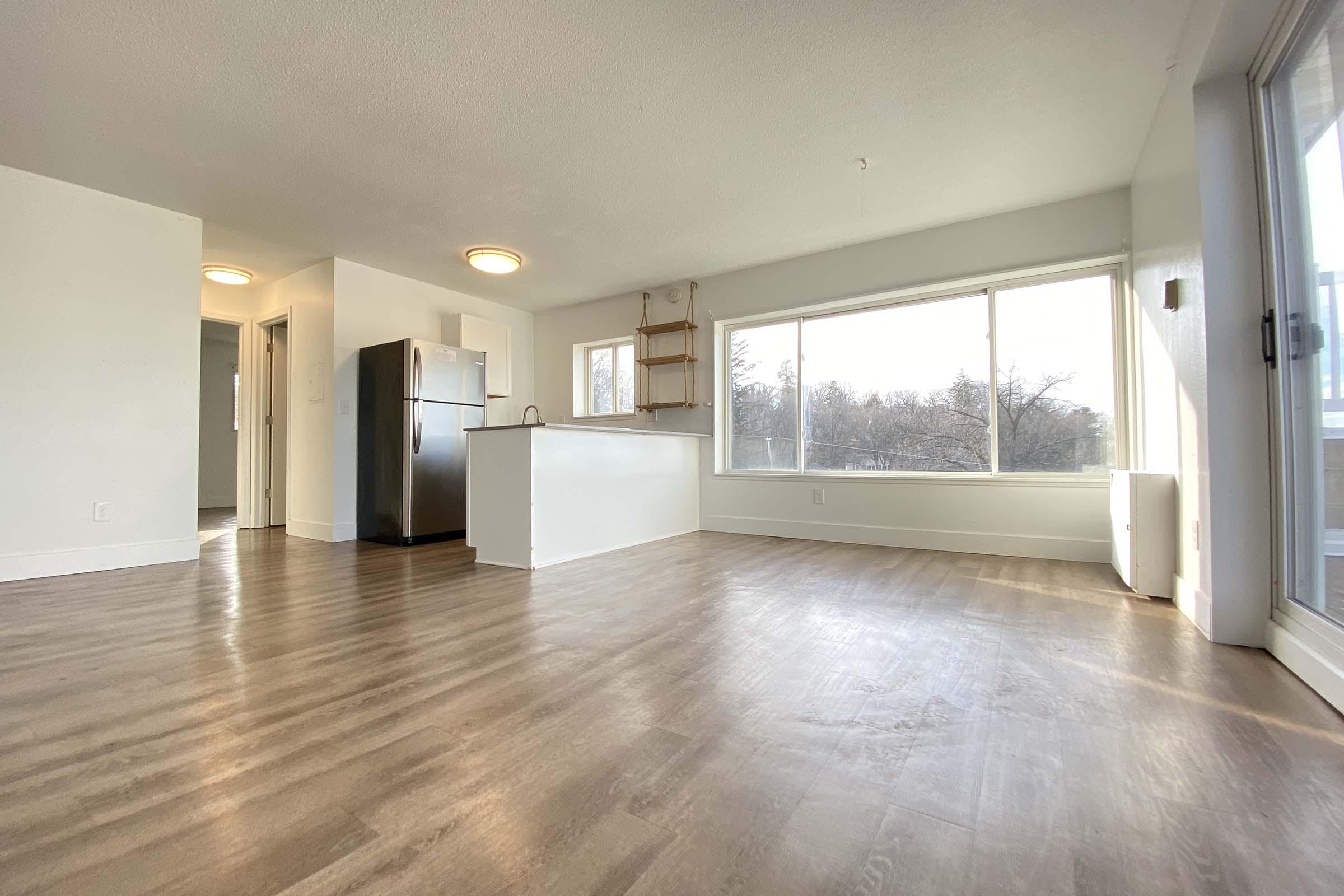 Modern, open-concept living space with large windows. Features a light-colored kitchen with a refrigerator, a spacious area with hardwood flooring, and natural light. The walls are painted in neutral tones, creating a bright and airy atmosphere.
