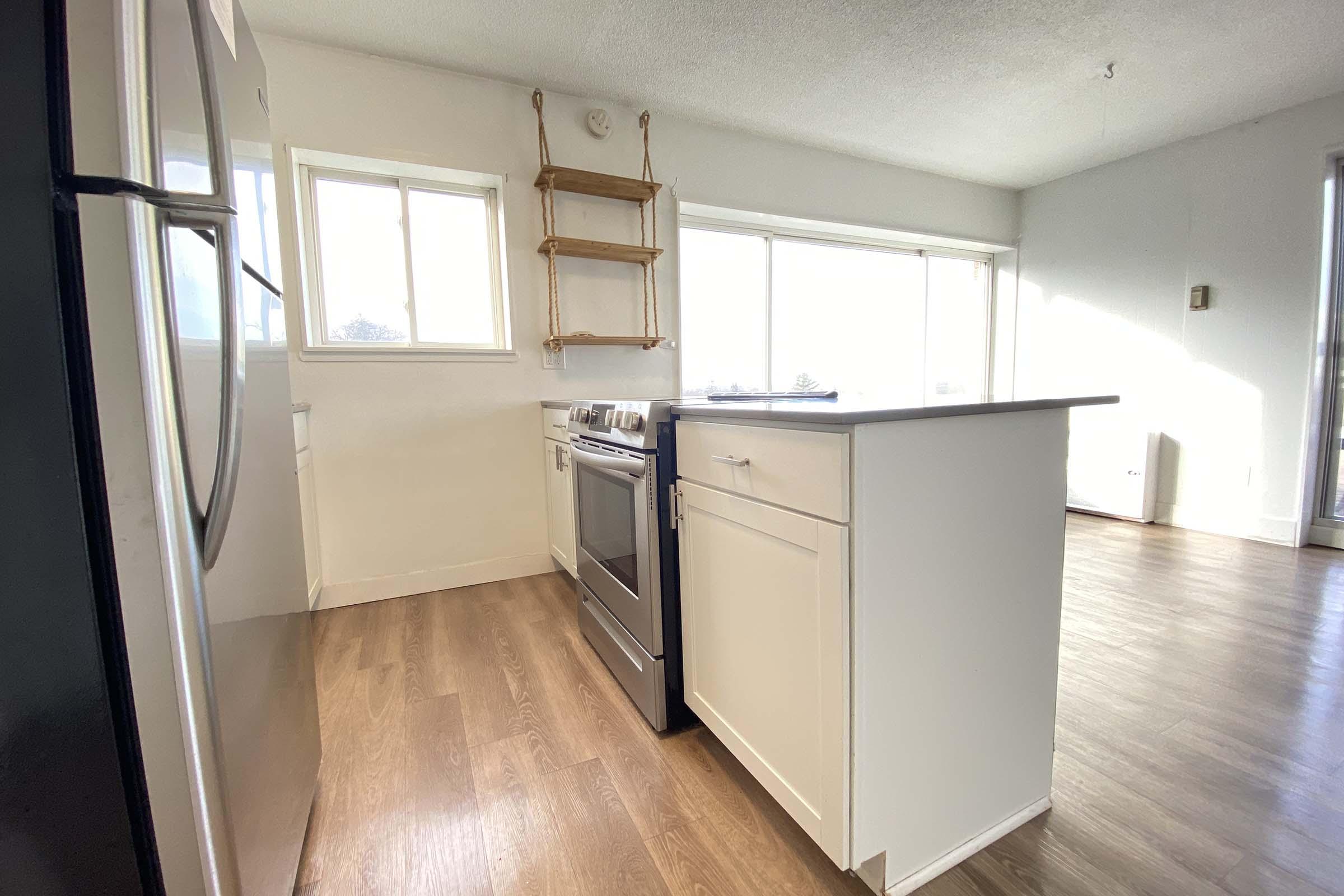 Modern kitchen with stainless steel appliances, including a stove and refrigerator. There’s a white island with a single drawer, wooden open shelves mounted on the wall, and large windows allowing natural light to fill the space. The flooring is light wood, creating a warm and inviting atmosphere.
