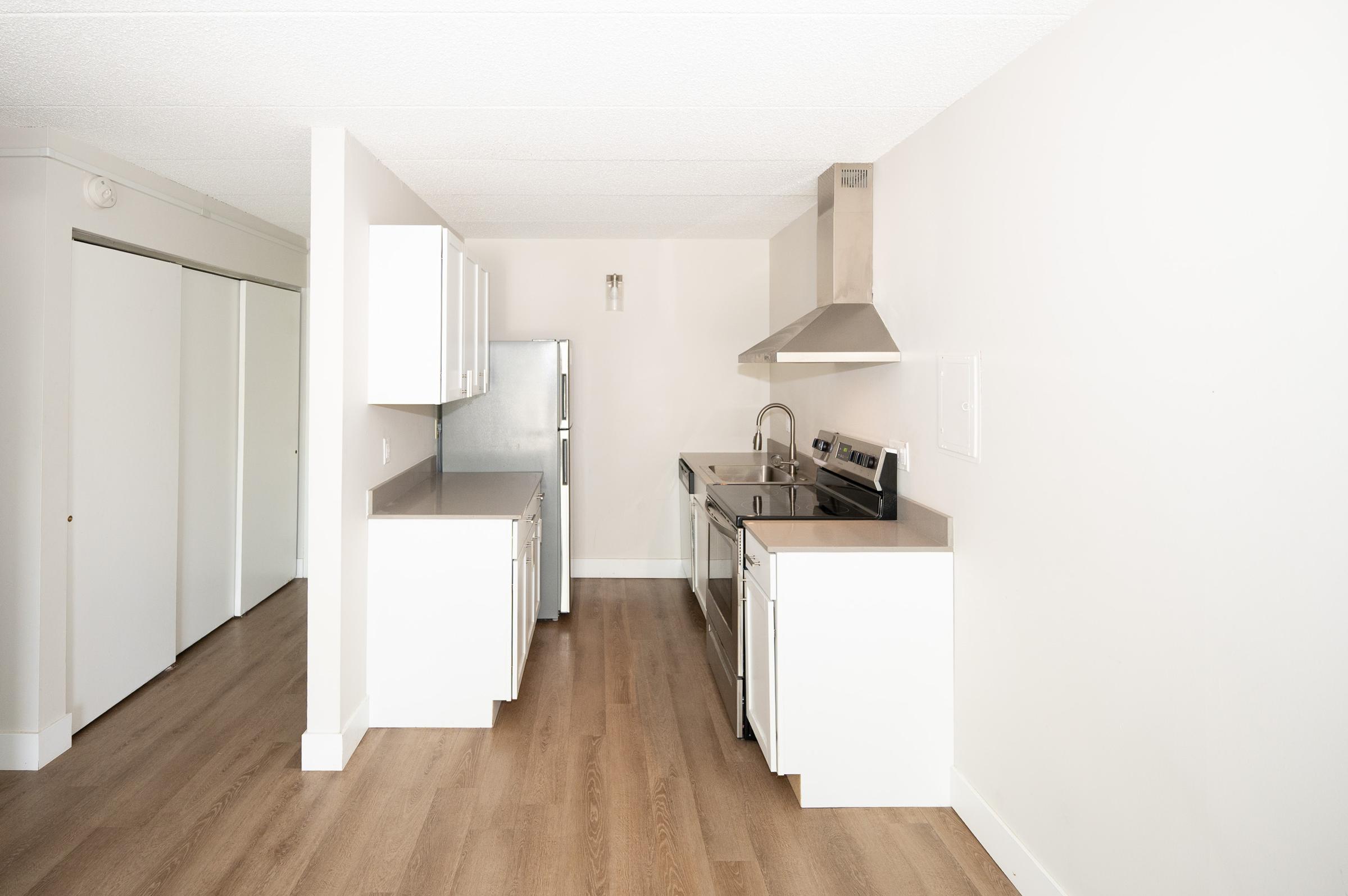 Modern kitchen featuring stainless steel appliances, including a stove and refrigerator, with white cabinetry and countertops. The space has a light, airy feel with wooden flooring and a minimalist design. A door leads to another room, suggesting an open-concept layout.