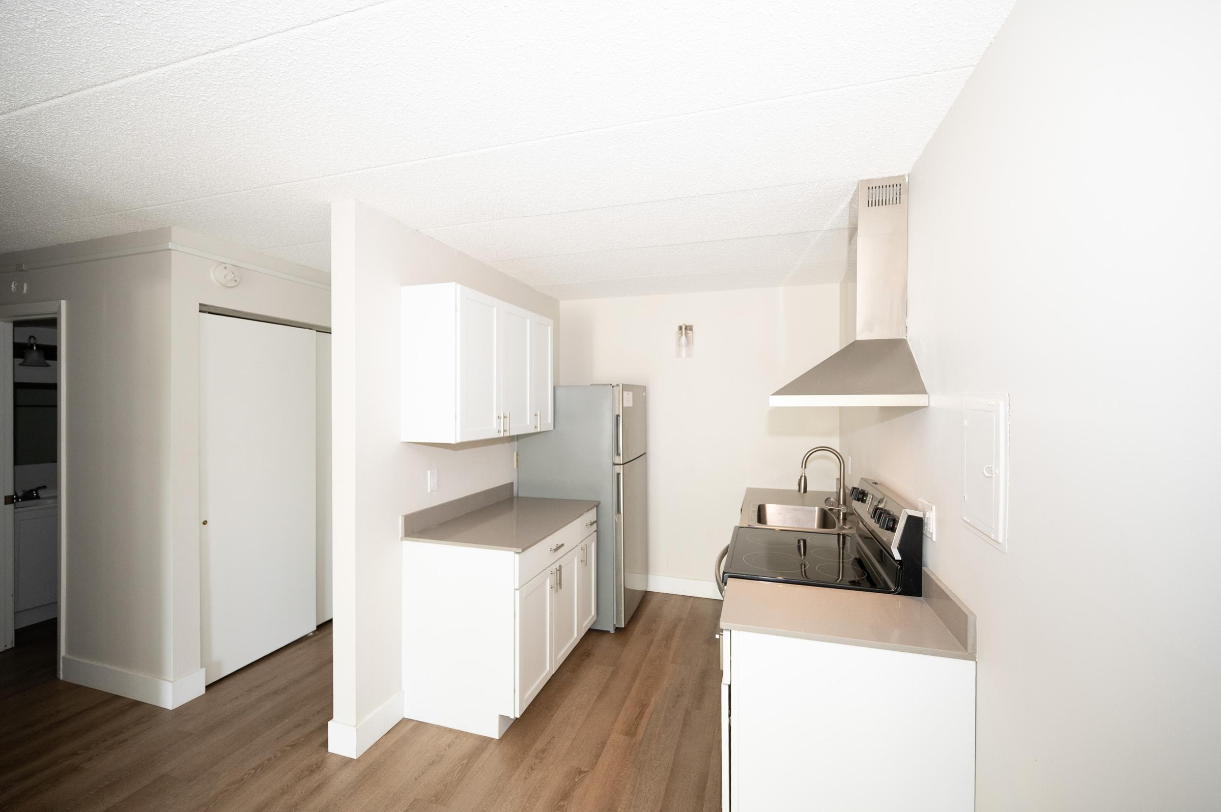 A modern kitchen featuring white cabinets, a stainless steel refrigerator, a double sink, and an electric stove. The room has light-colored walls and a light wood floor, with a door leading to another space visible in the background. Bright and spacious with ample storage.