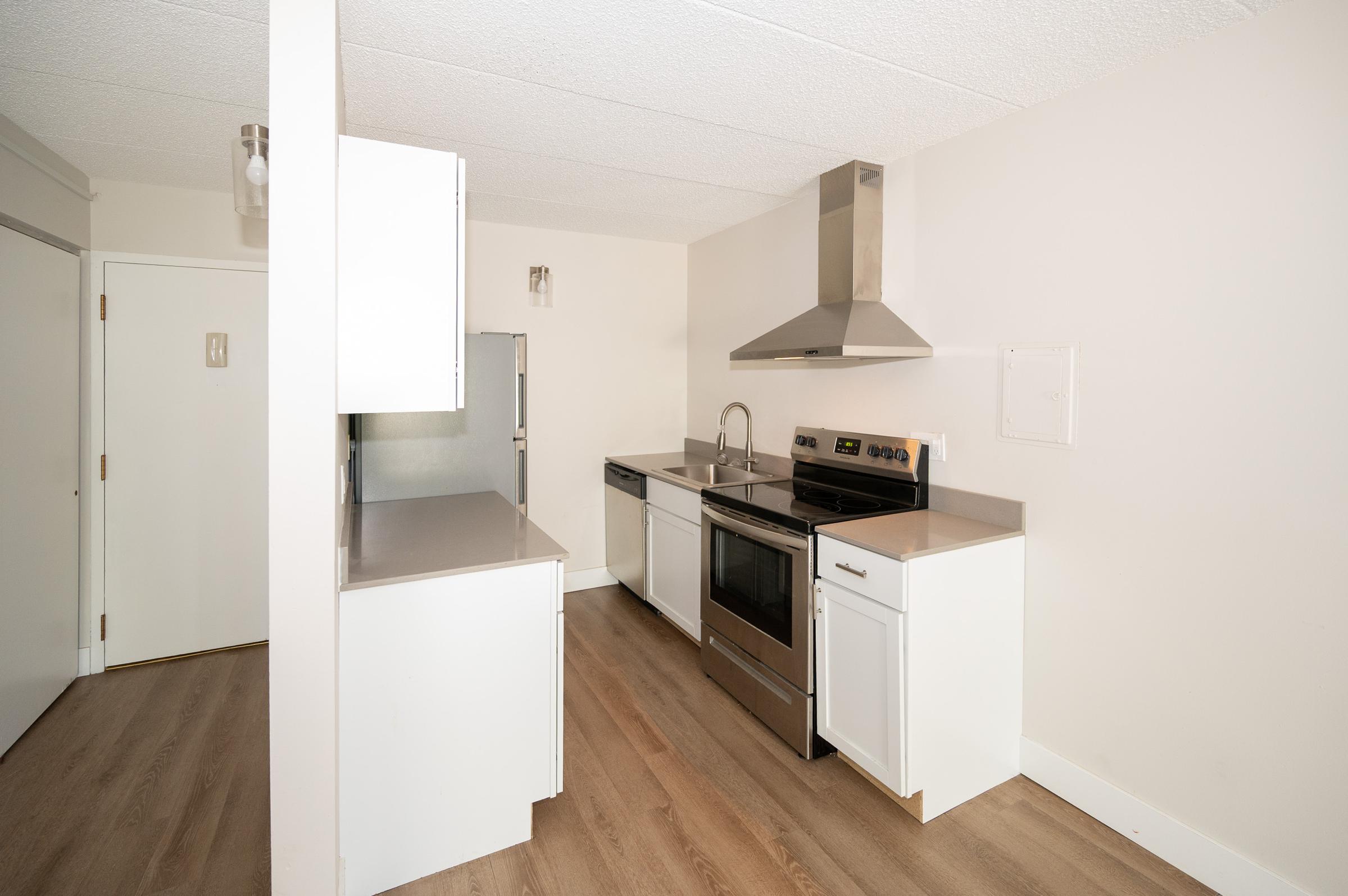 Modern kitchen with stainless steel appliances, including a stovetop, oven, and refrigerator. The cabinetry is white with a sleek design, and the countertops are gray. The room features light wood flooring and neutral wall colors, creating a bright and clean atmosphere.