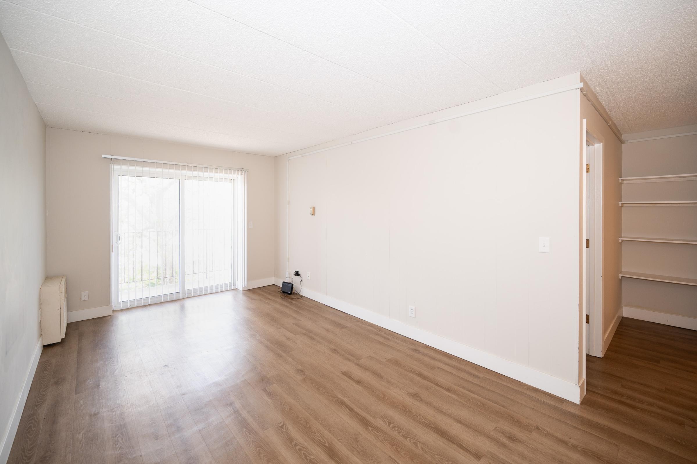 A spacious, empty room with light-colored walls and a wooden floor. There is a sliding glass door leading to an outdoor area and a small shelf along one wall. The room appears bright and well-lit, with no furniture or decorations present.