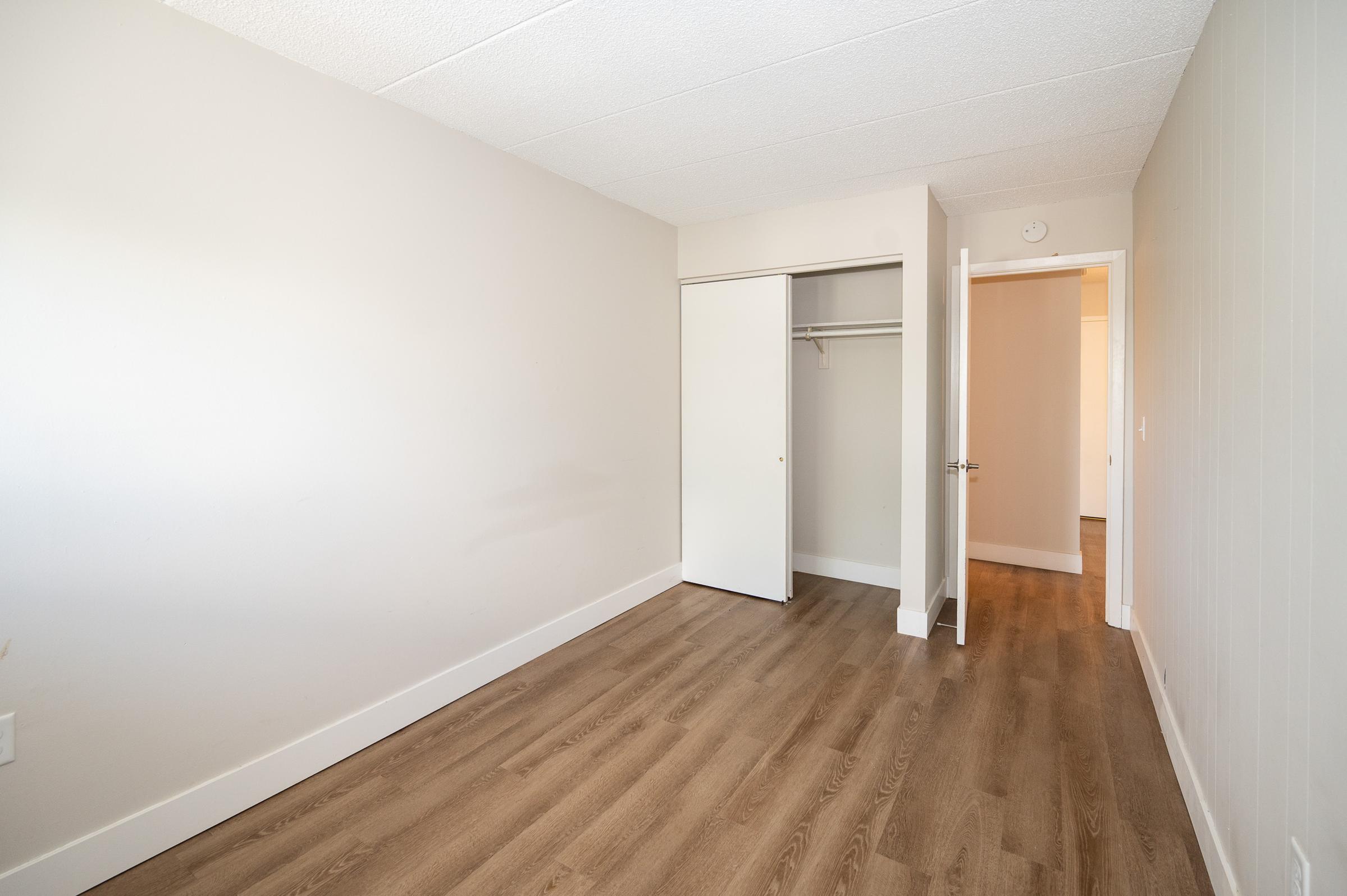 A simple, empty room with light-colored walls and a hardwood floor. There is a closet with a white sliding door on one side and an open doorway leading to another room on the opposite side. The space is well-lit and features a clean, minimalist design without furniture.