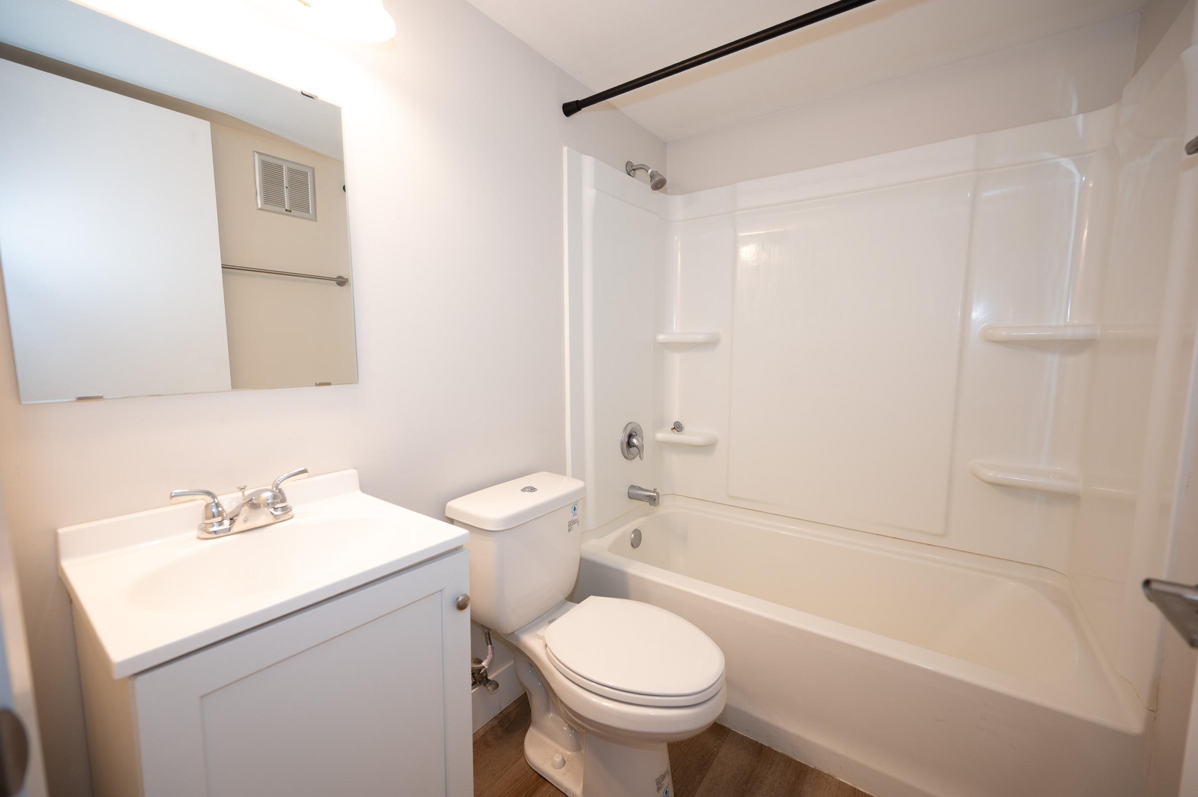 A clean bathroom featuring a white bathtub with a shower, a modern toilet, and a sink with a cabinet underneath. A mirror is mounted above the sink. The walls are painted white, and the flooring appears to be a light wood look. Soft lighting enhances the bright, open feel of the space.