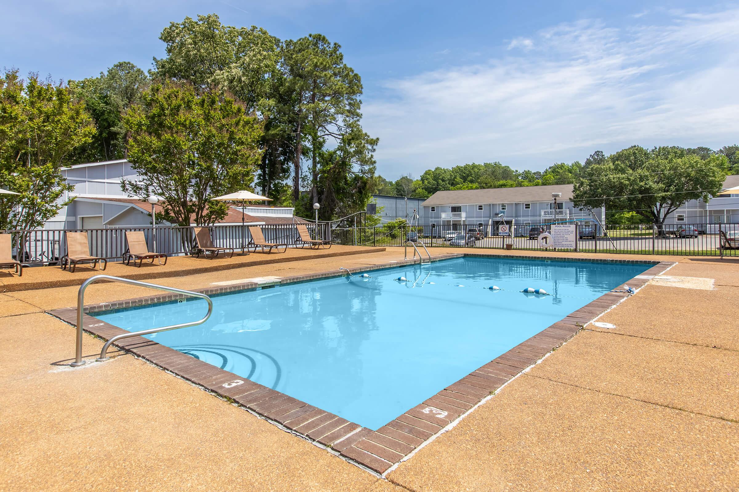A bright outdoor swimming pool surrounded by a tan concrete deck. There are a few lounge chairs positioned nearby, with green trees and a clear blue sky in the background. Residential buildings are visible in the distance, creating a relaxing atmosphere.