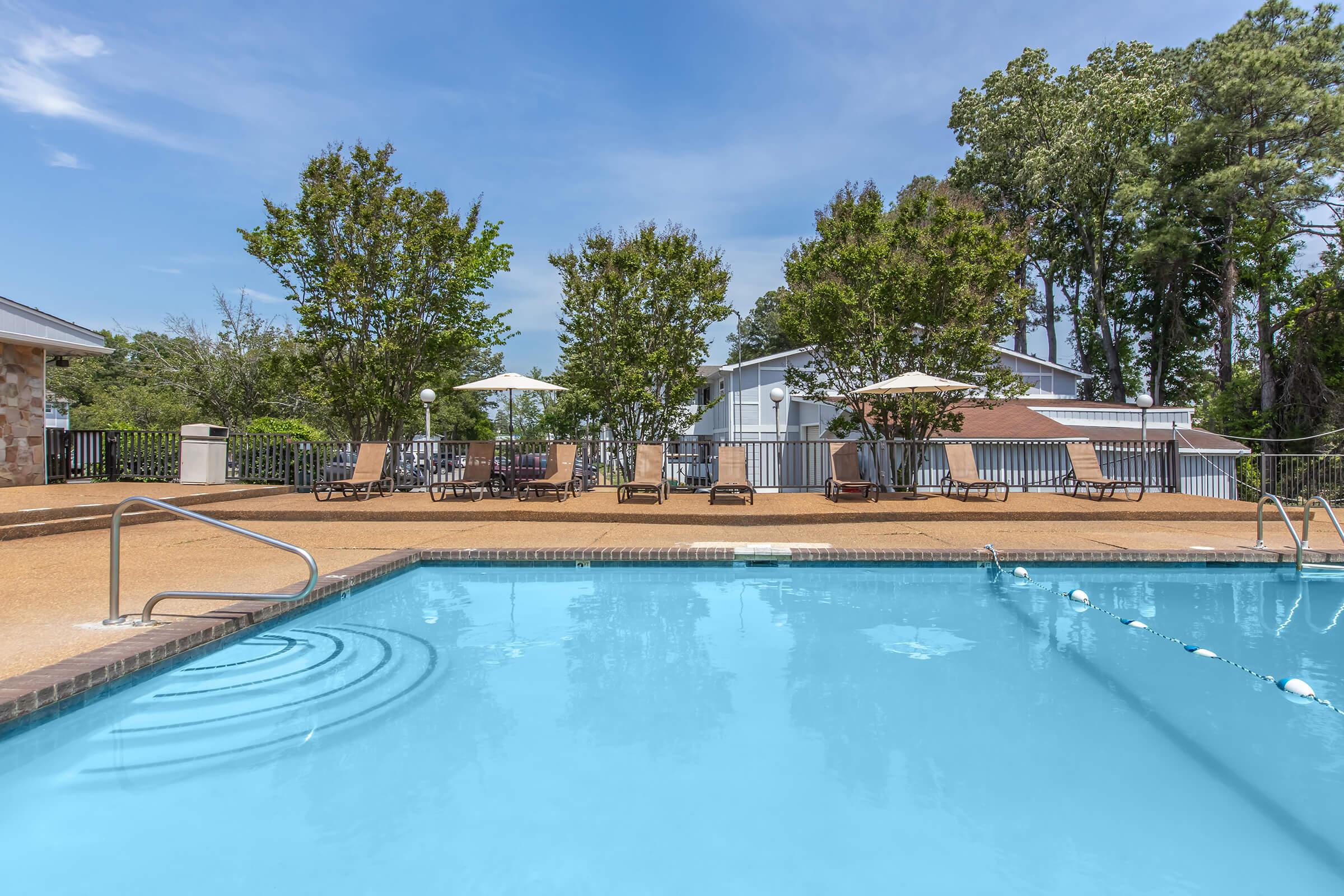 A clear blue swimming pool with gentle ripples, surrounded by lounge chairs and umbrellas. Lush green trees provide shade in the background, and a building is partially visible behind the pool area, under a bright blue sky with a few clouds. The scene suggests a relaxing outdoor environment.