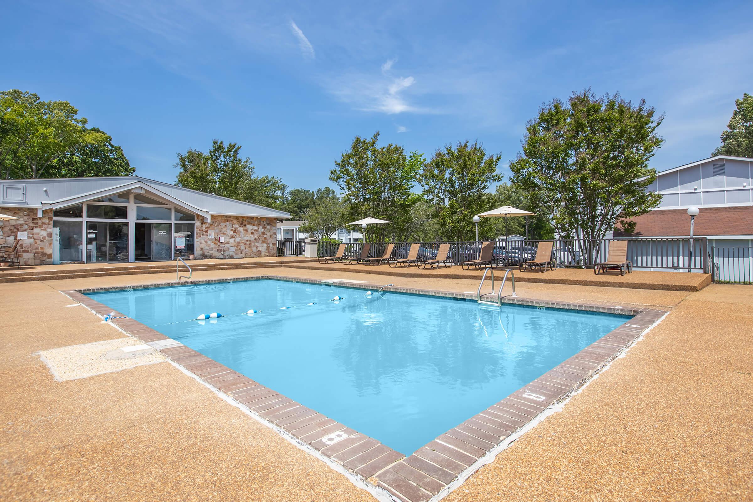 A clear blue swimming pool surrounded by a tan deck. Comfortable lounge chairs are lined up along the poolside, and shaded umbrellas are positioned for relaxation. In the background, there are trees and a building with large windows, creating a serene and inviting atmosphere.