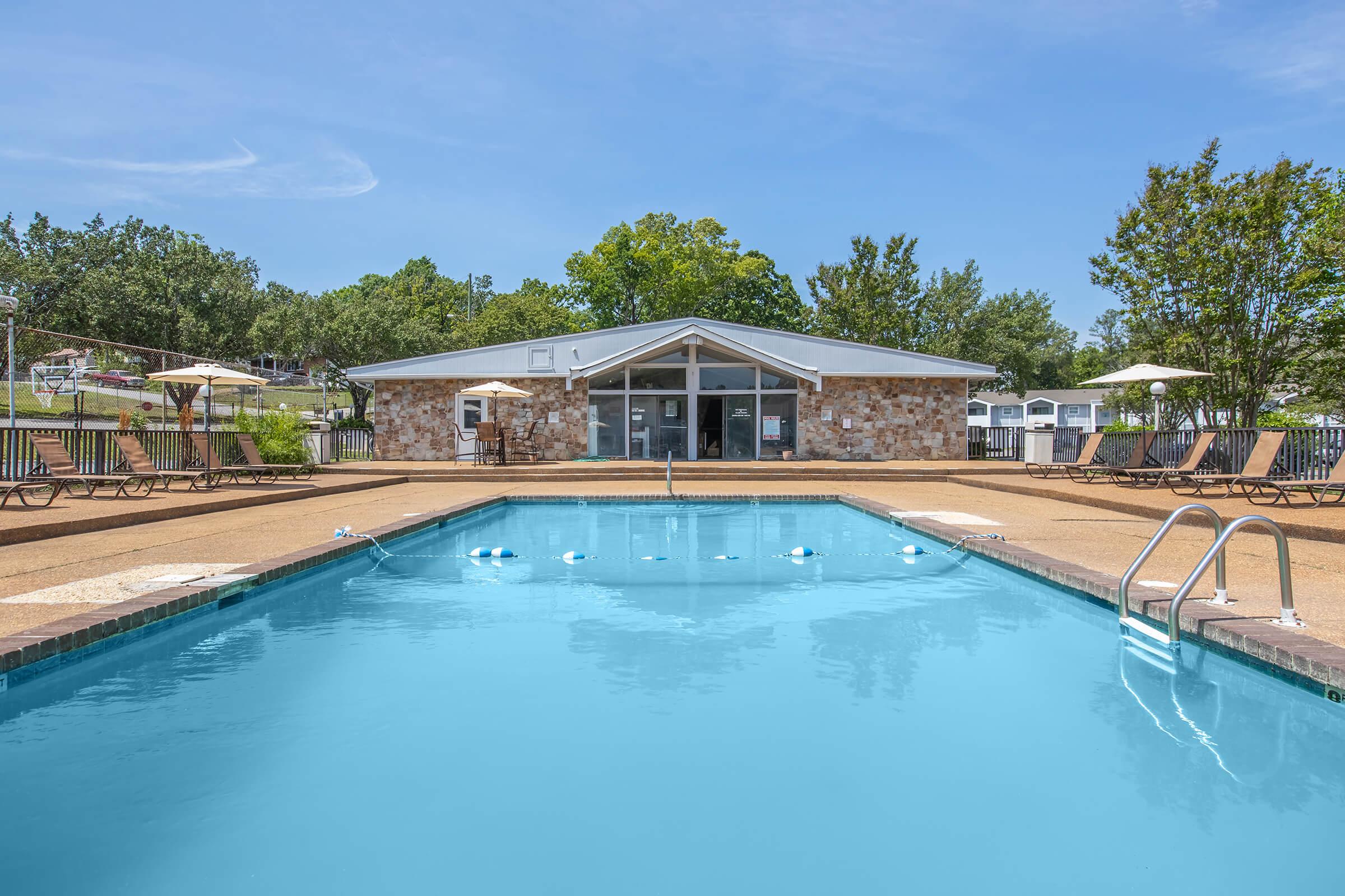 A clean, bright swimming pool with a clear blue surface, surrounded by lounge chairs and umbrellas. In the background, there is a modern building with a stone façade and large windows, set against a backdrop of trees and a clear blue sky.