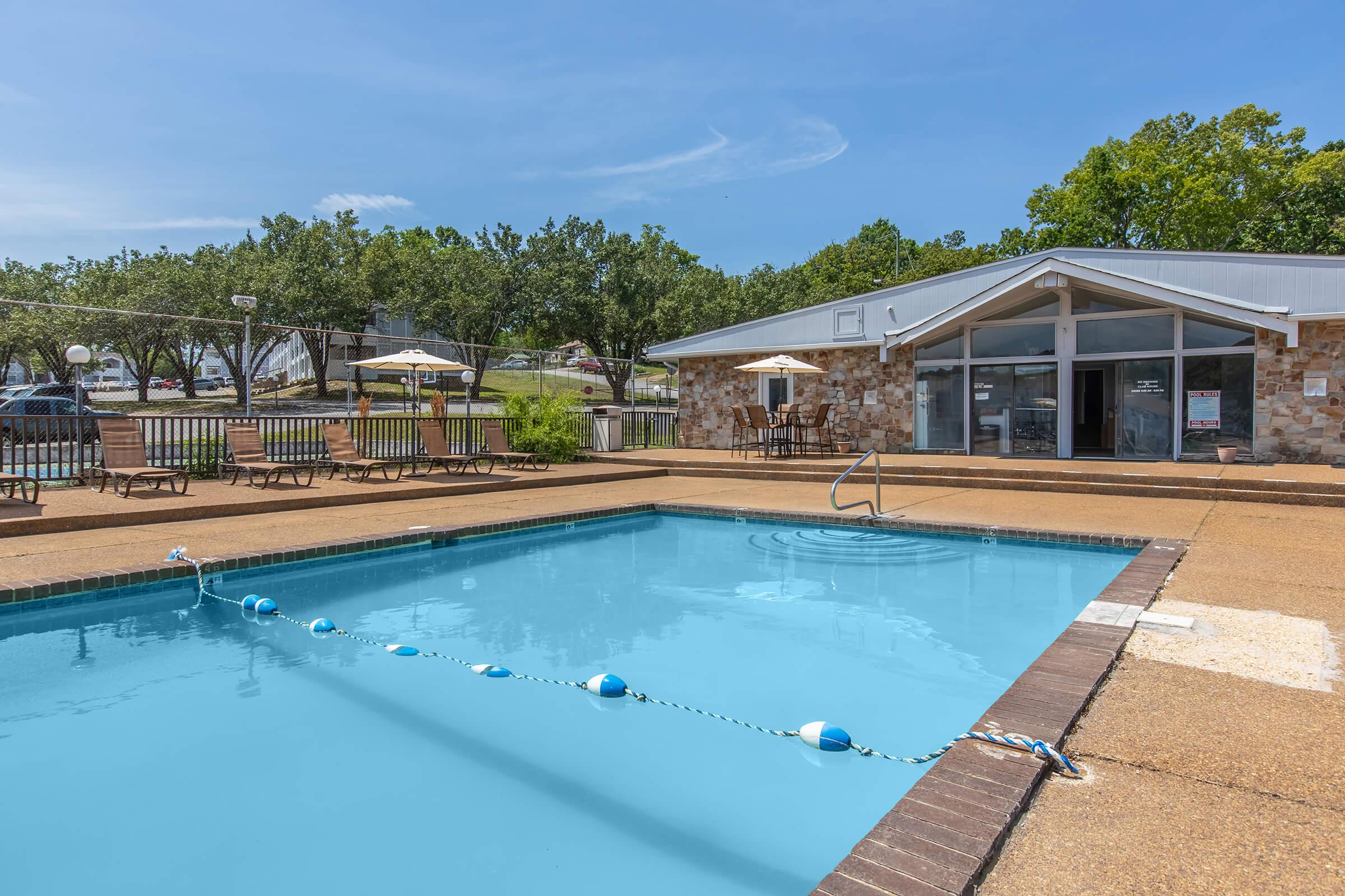 A clear blue swimming pool surrounded by a concrete deck, with lounge chairs on one side and a small building in the background. The area is sunny, with a few trees and patio umbrellas visible. A safety line with buoys is placed in the pool.