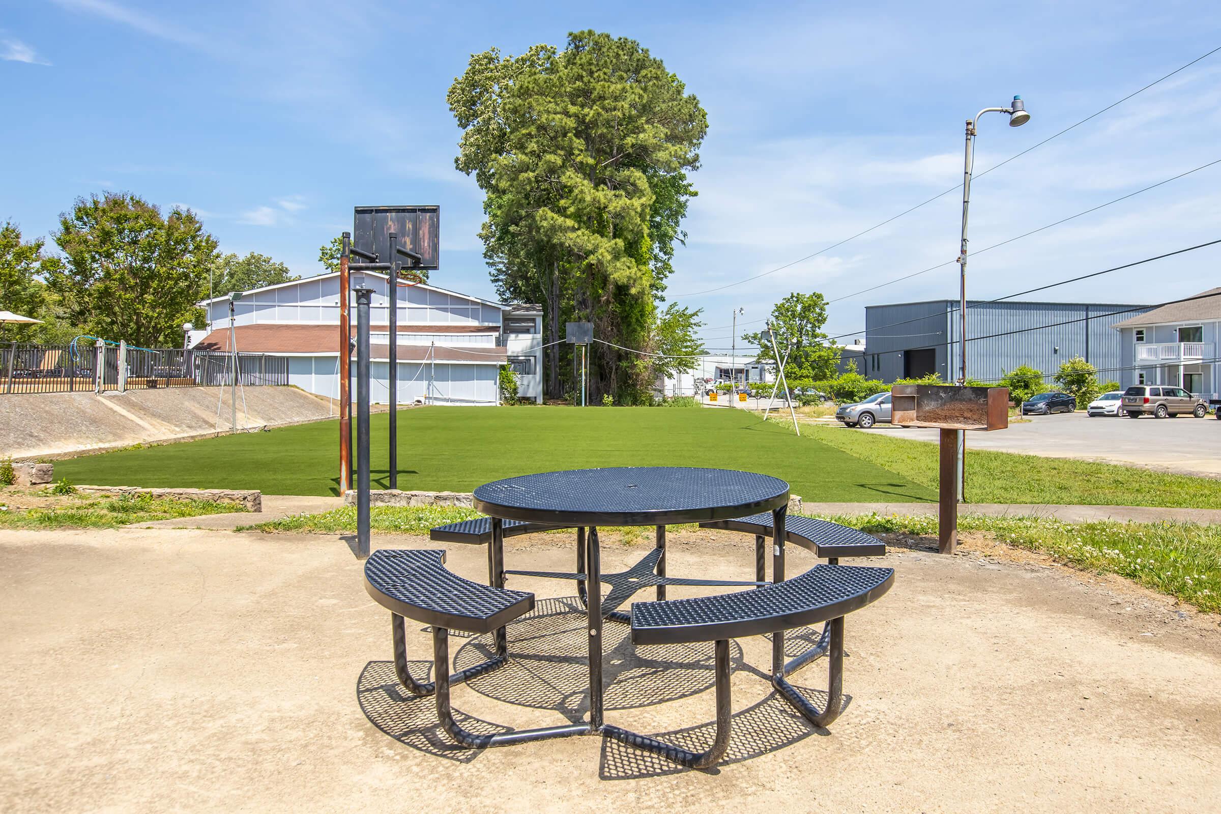 A circular metal picnic table with attached benches sits on a concrete surface in a park. In the background, a basketball hoop is visible, along with a grassy area and trees. A building can be seen further back, and a light post stands nearby under a clear blue sky.