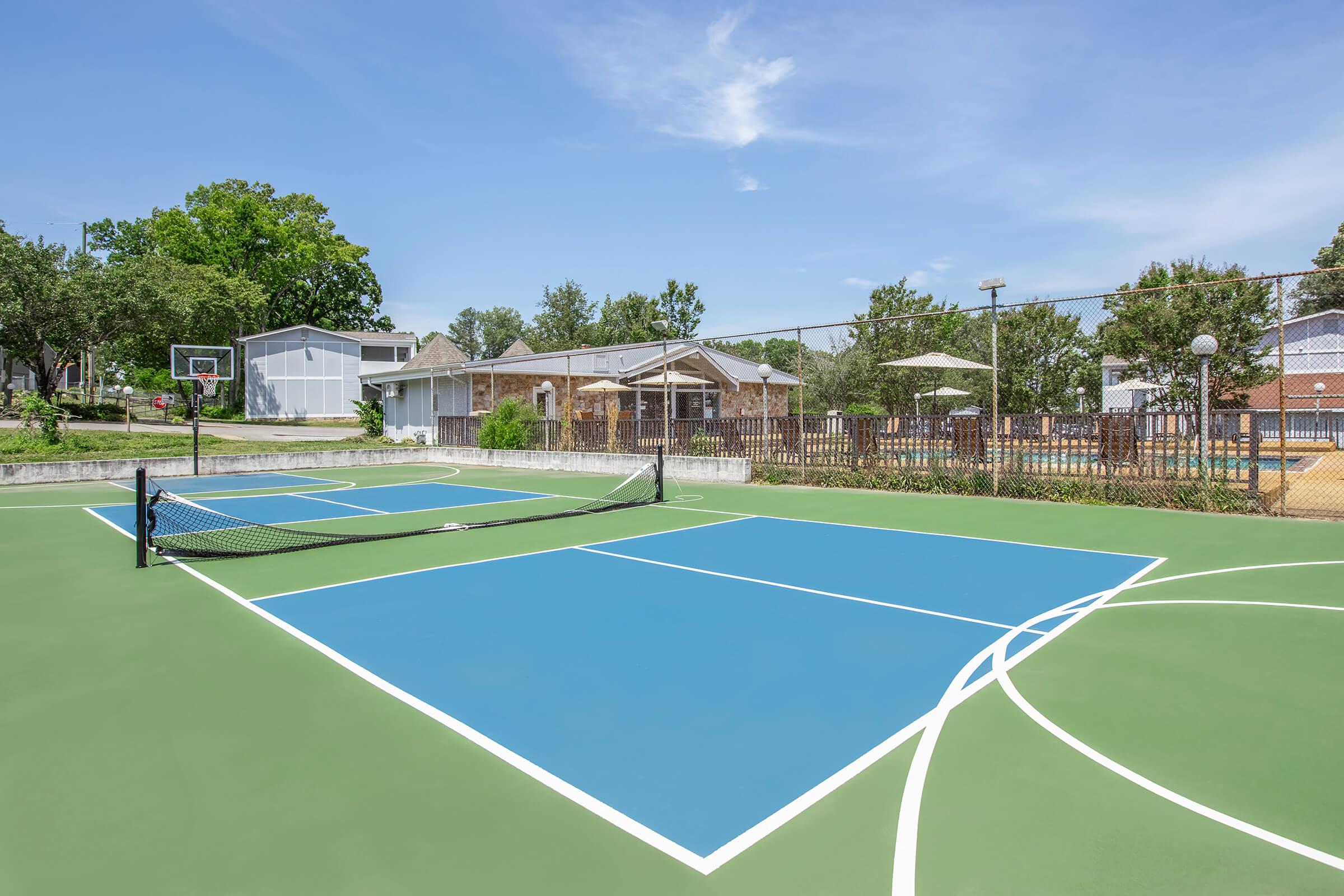 A view of a multi-sport court featuring blue tennis courts and a basketball hoop in a sunny outdoor setting. Surrounding the courts are trees and residential buildings, with a clear blue sky overhead. The area is well-maintained and ideal for outdoor sports activities.