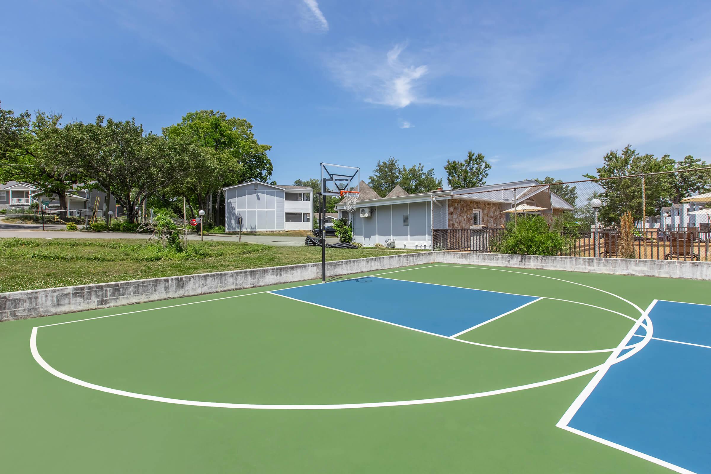 A basketball court featuring vibrant green and blue markings, with a hoop but no players. In the background, there are residential houses and trees under a clear blue sky, creating a peaceful and inviting atmosphere. The court is well-maintained and surrounded by grassy areas.