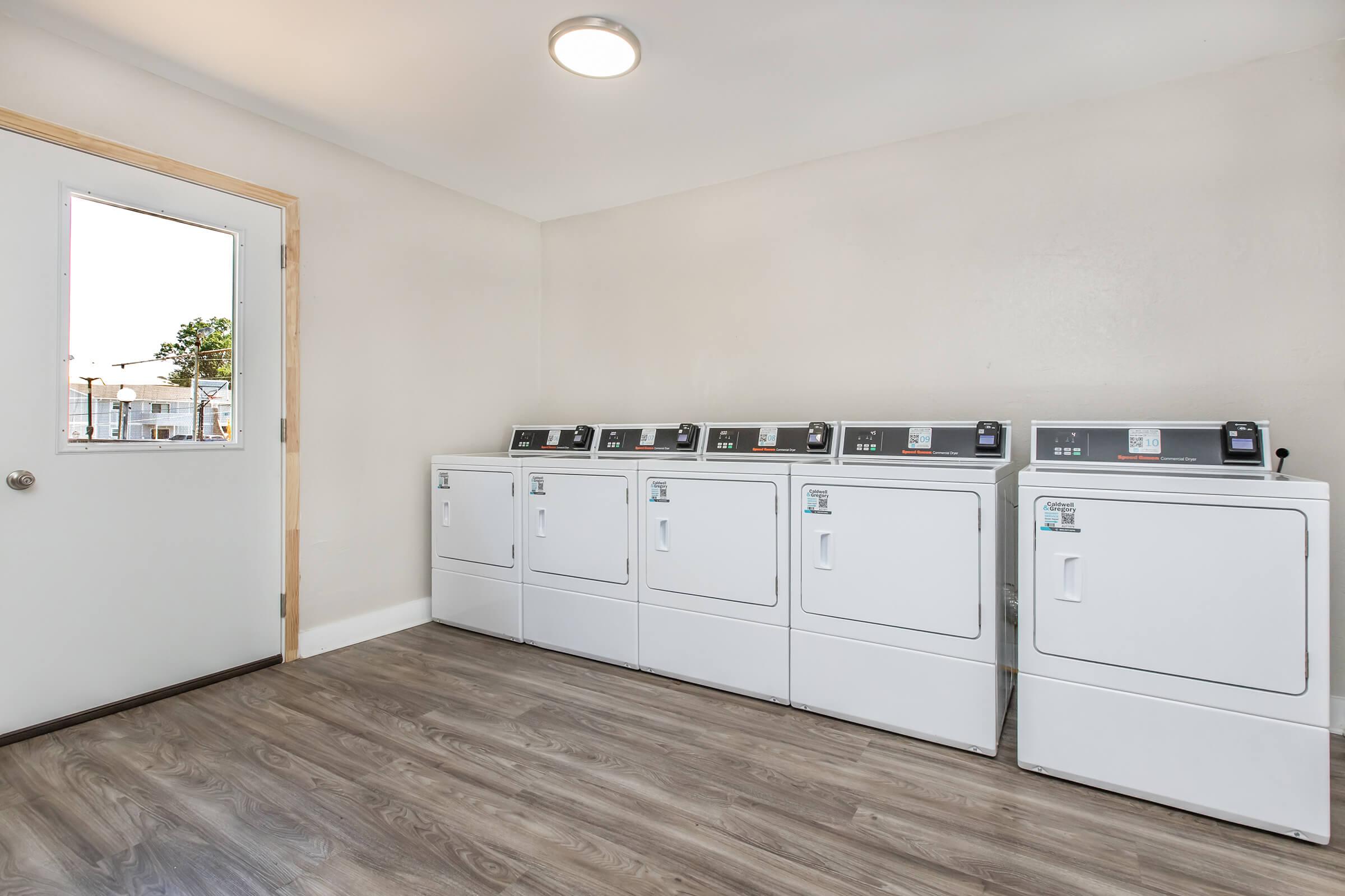 A clean and bright laundry room featuring five white washing machines lined up against a wall. There is a door leading outside, and the room has a light wooden floor. The walls are painted in a light color, creating a spacious and inviting atmosphere.