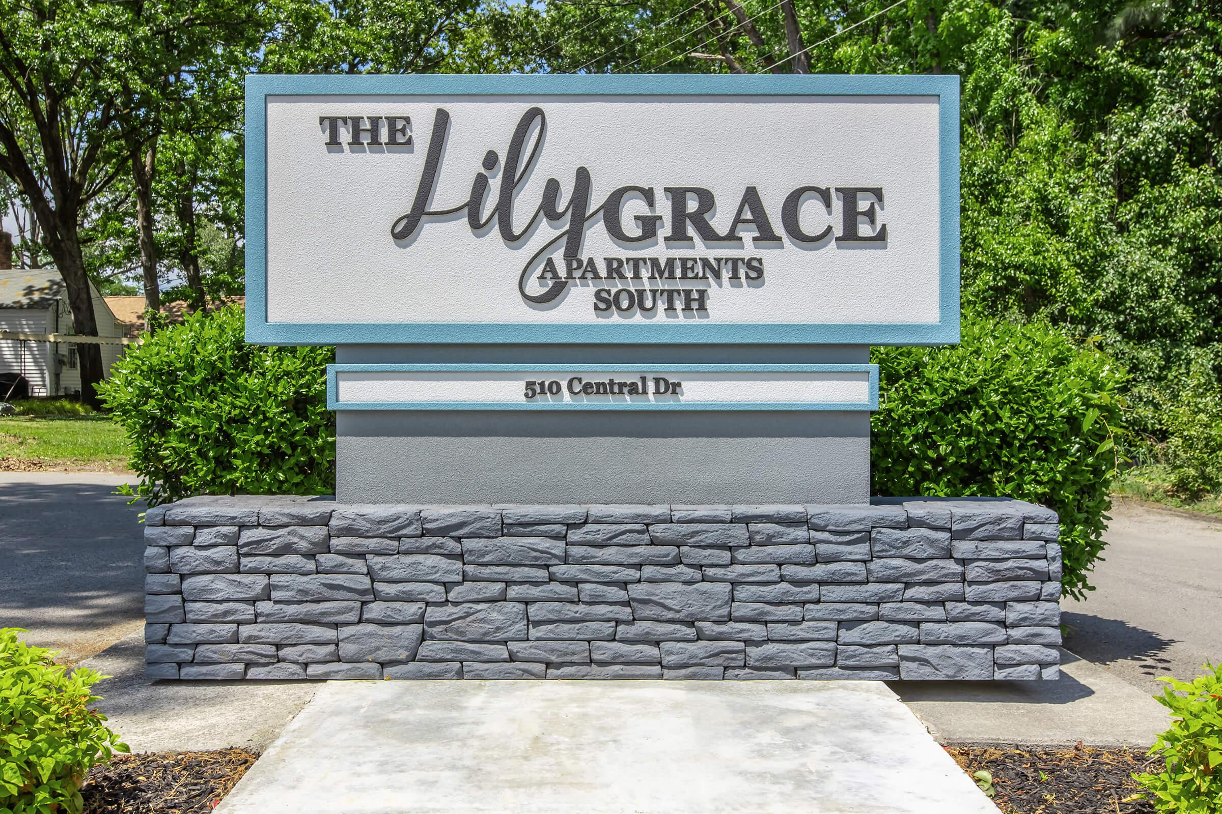 Sign for The Lily Grace Apartments South, located at 510 Central Dr. The sign features a modern design with the name prominently displayed in large letters against a light-colored background, bordered by a stone base and surrounded by greenery.