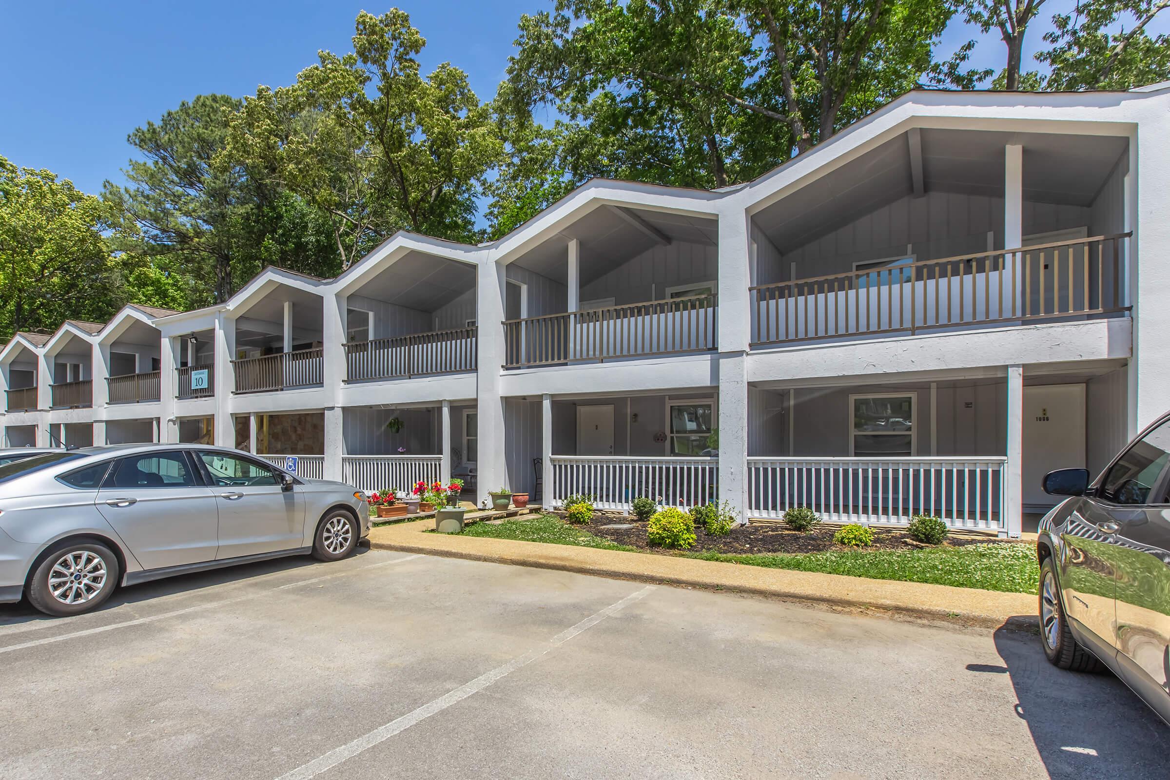 A row of white, multi-level building units with unique, peaked roofs. Each unit has a balcony with railings. The area is landscaped with small shrubs and flowers, and there are parked cars in the foreground on a paved lot. The setting is sunny with clear blue skies and trees in the background.