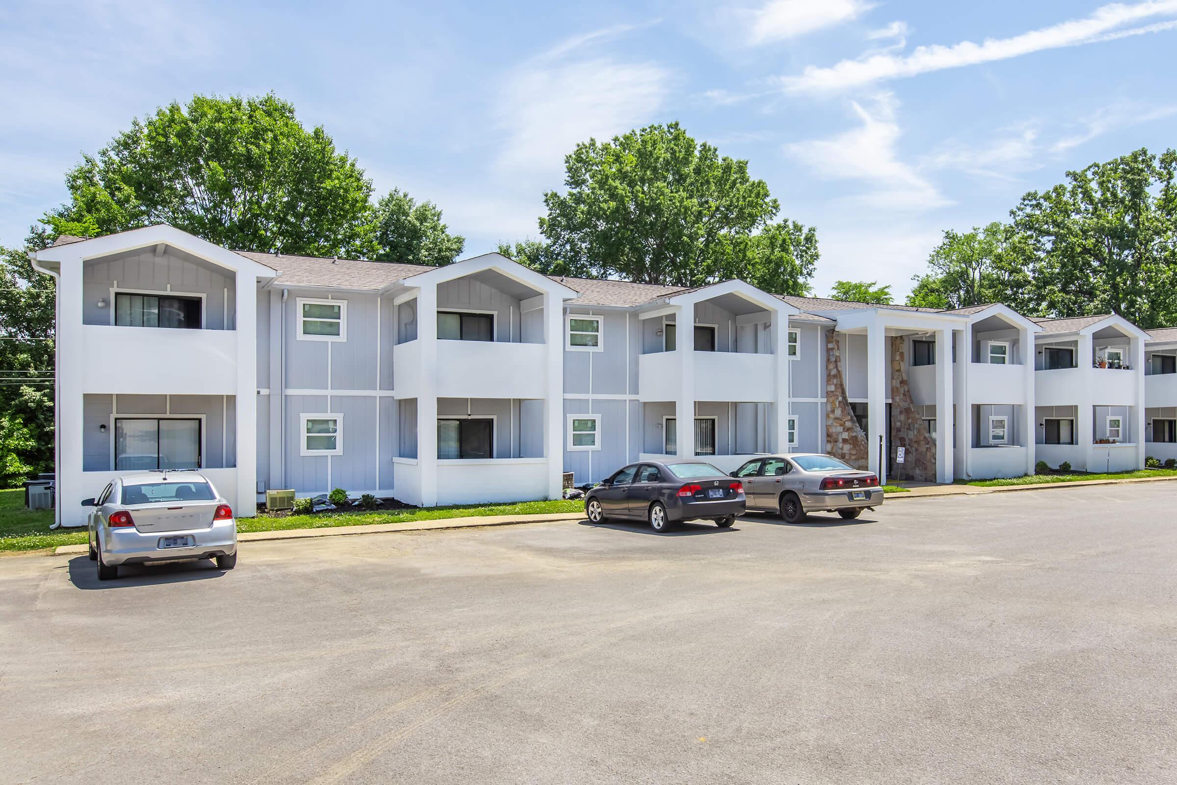 A modern low-rise apartment building featuring multiple white and gray units with pitched roofs. The building is set against a clear blue sky and surrounded by green trees. There are several parked cars in the foreground, and the parking lot is paved with asphalt.