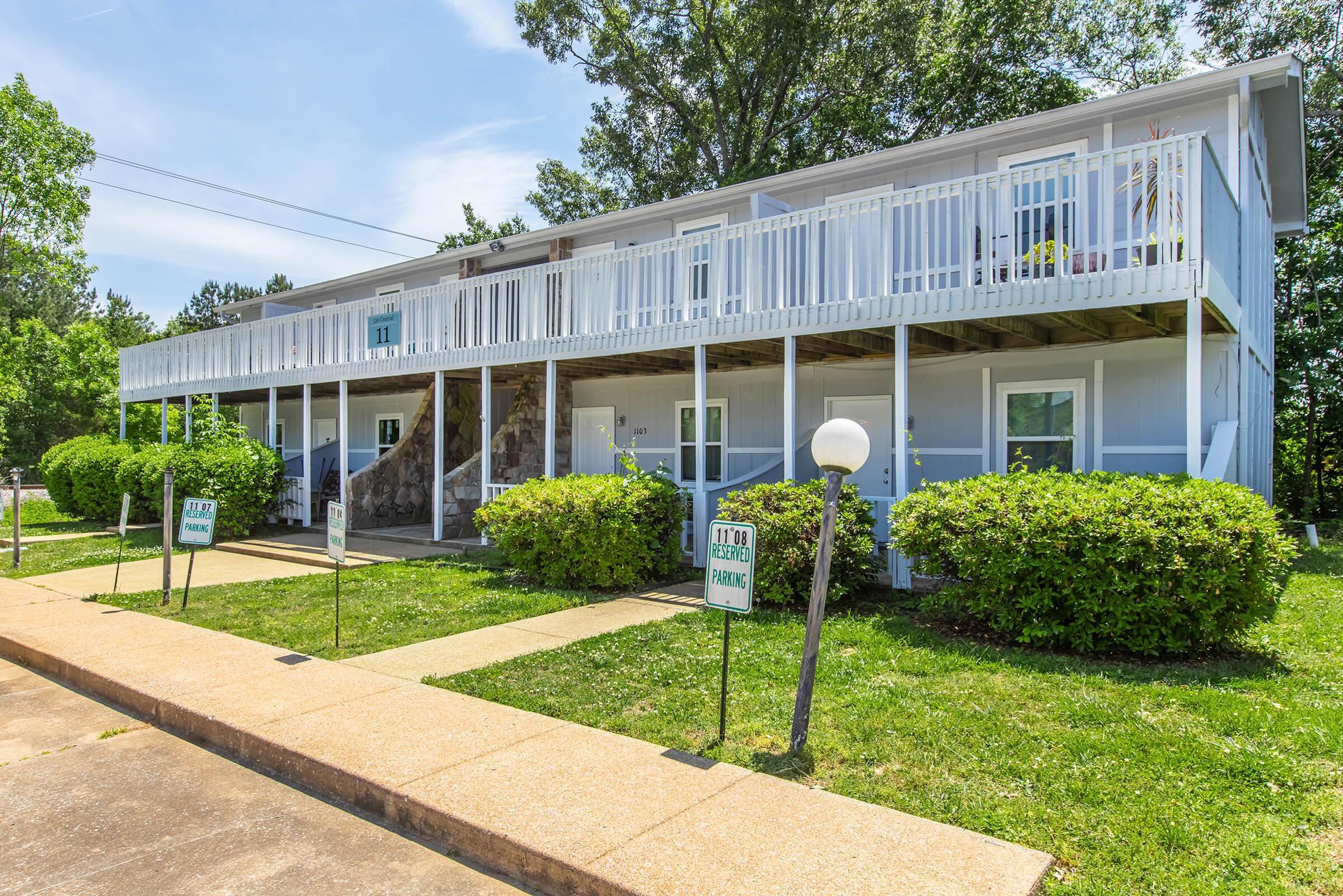 Two-story residential building with white siding and a covered walkway on the ground floor. Green bushes line the front, and there are signs for parking. The area is sunny with trees in the background and a clear blue sky.