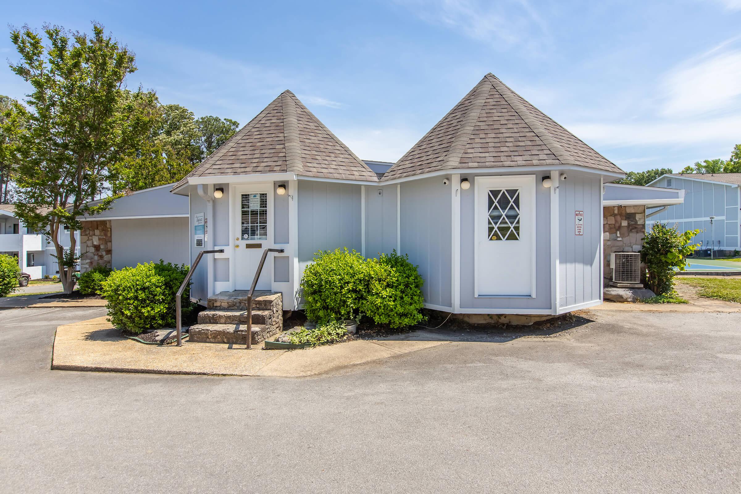 Two small, charming buildings with pointed roofs, surrounded by lush greenery and concrete pathways. The structures have large windows and light-colored exteriors, situated in a sunny outdoor setting with a clear blue sky.