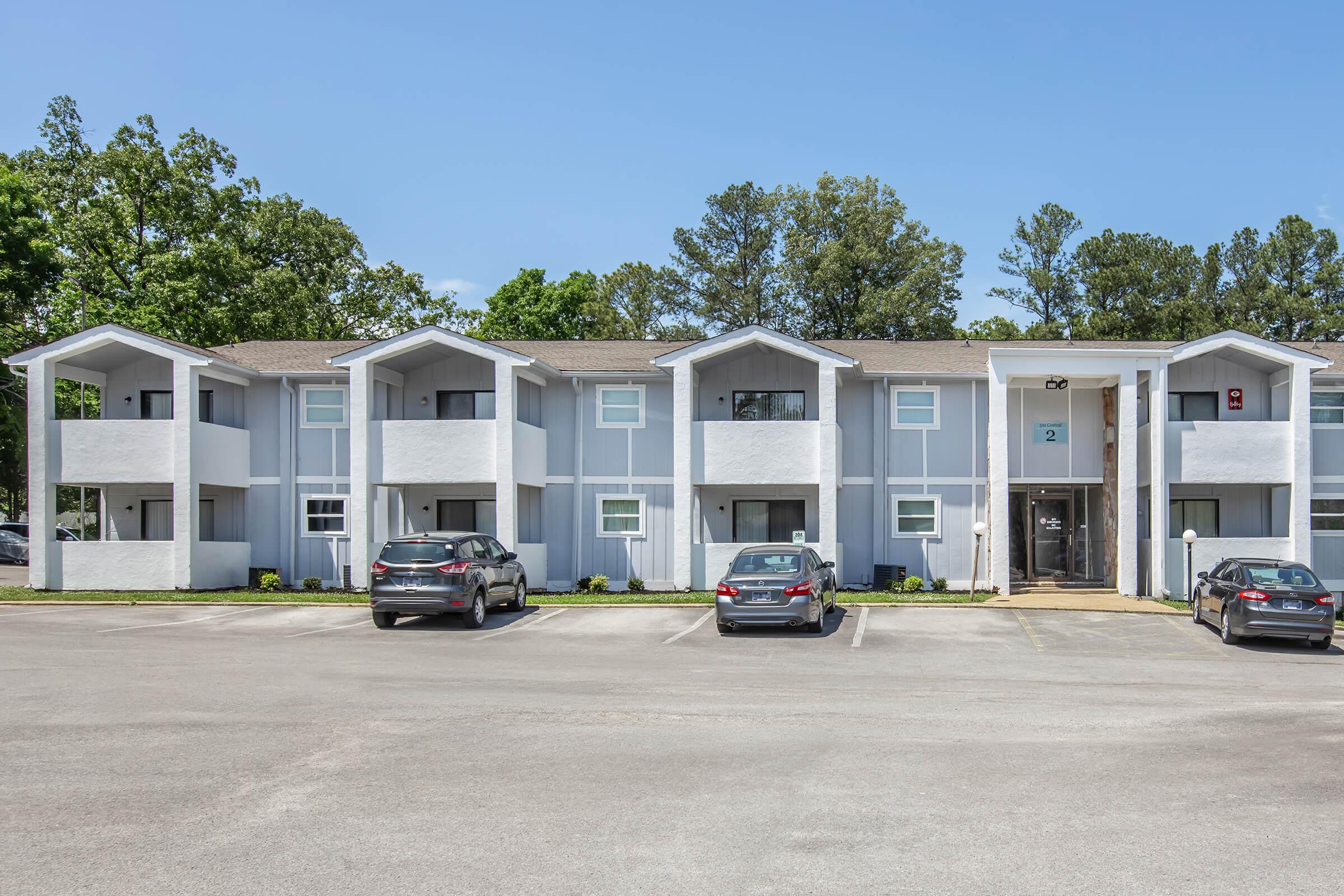 A modern three-story apartment building featuring a light gray exterior with multiple units, each having distinctive peaked roofs. The facade includes windows and access doors. Several parked cars are visible in the foreground, with a clear blue sky and green trees in the background.