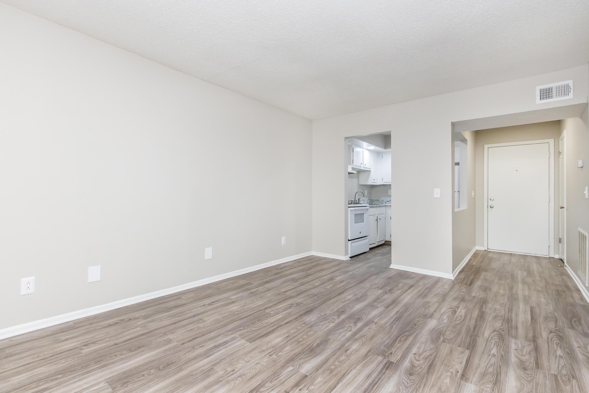 A spacious, empty living room with light-colored walls and laminate flooring. A doorway leads to a kitchen area with white cabinetry and appliances visible in the background. Natural light fills the room, emphasizing the clean and minimalist layout. A front door is seen on one side, with ample wall space for decorating.