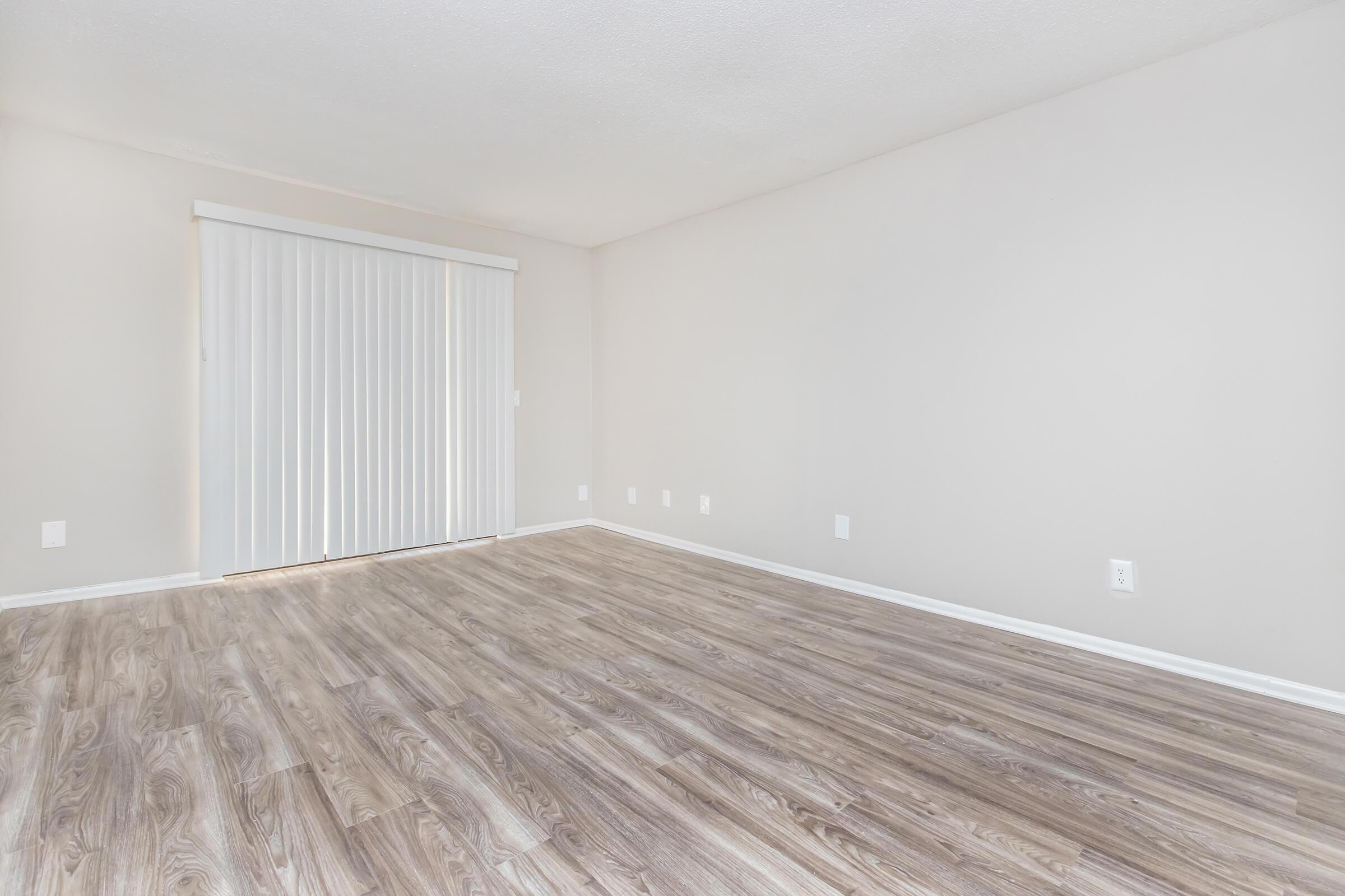 A vacant room with light gray walls and a wood-like laminate floor. There is a sliding door with vertical blinds on one side, allowing natural light to enter. The space is uncluttered and minimalistic, ideal for various uses or decorating.