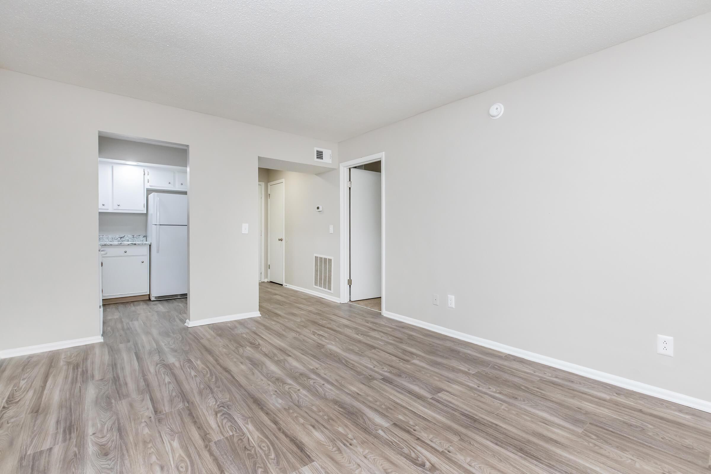 A spacious, empty room with light-colored walls and a hardwood floor. There is an open entryway leading to a small kitchen area with white cabinets and appliances visible. Two doorways lead to other rooms, and the overall atmosphere is bright and neutral.