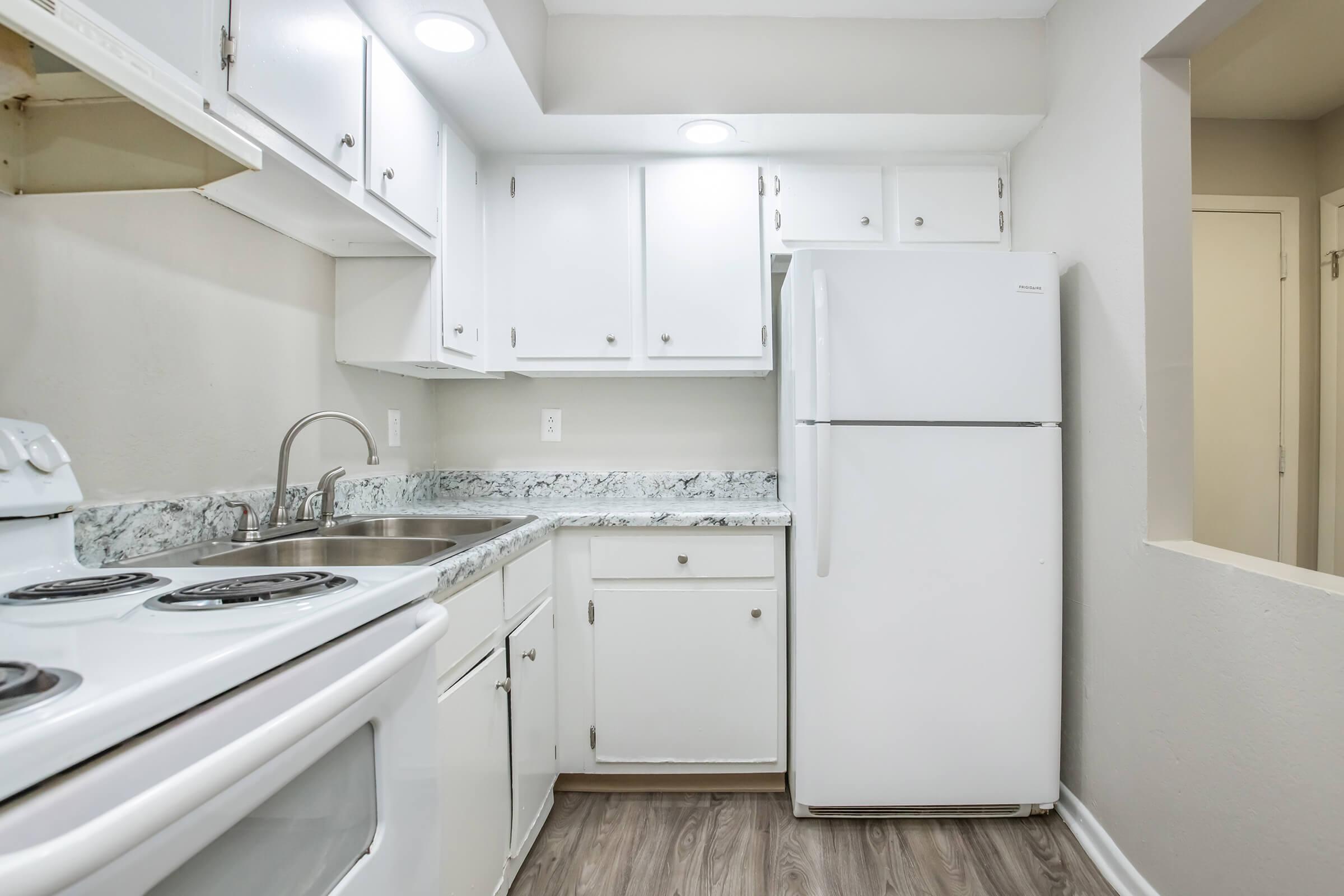 A small, modern kitchen featuring white cabinets, a stainless steel sink, a white refrigerator, and an electric stove with a four-burner cooktop. The room has light-colored walls, a granite countertop, and bright overhead lighting, creating a clean and inviting atmosphere.
