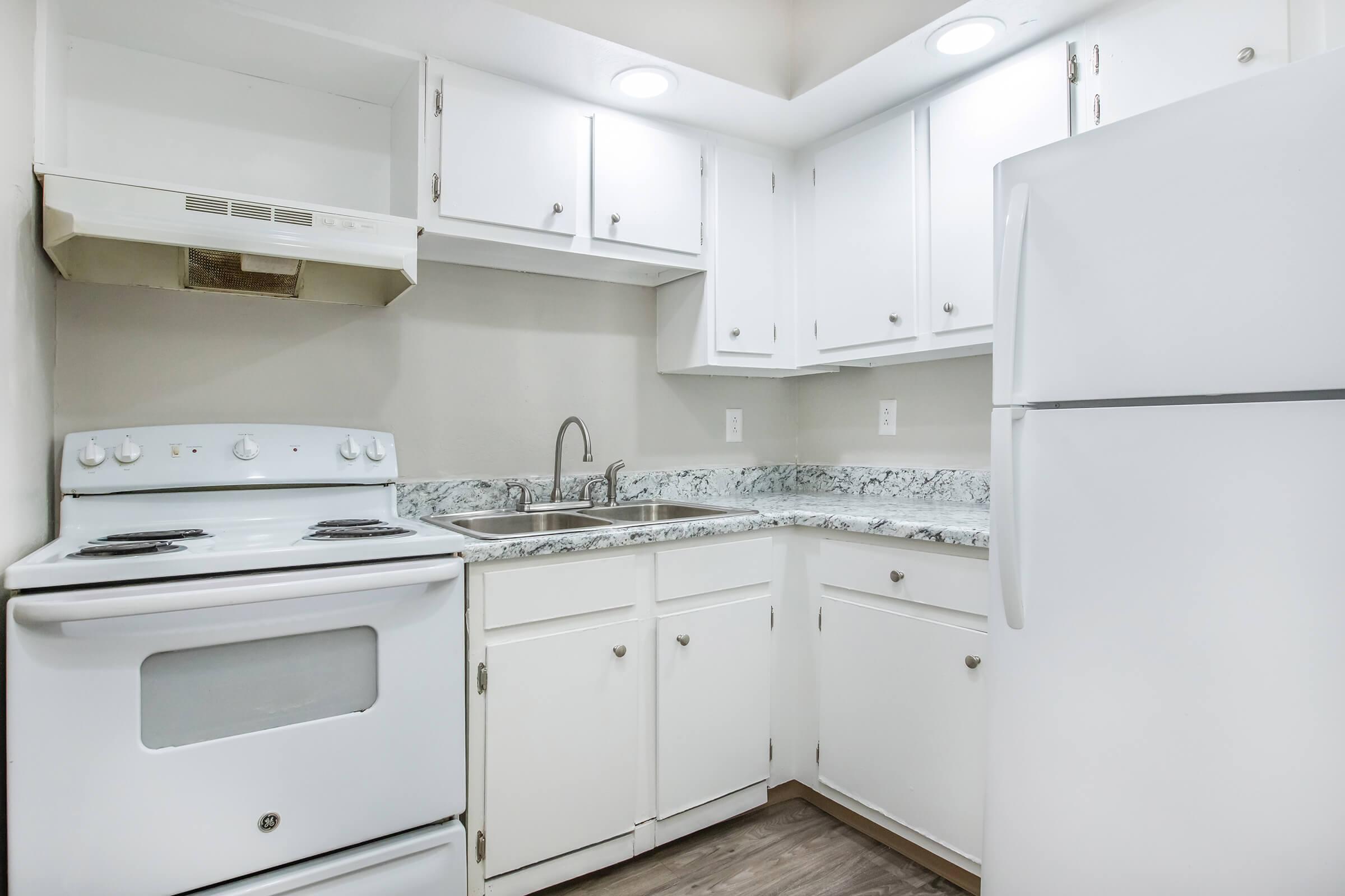 A modern kitchen featuring white cabinetry, a white stove, and a refrigerator. The countertops are speckled with gray and white patterns, and there's a small sink with a single faucet. The walls are light-colored, and the space is well-lit with overhead lighting.