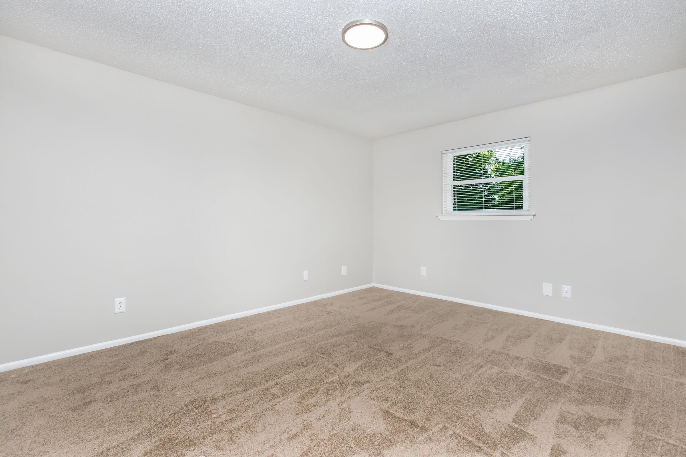 Empty room with light-colored walls and a light brown carpet. A window with white blinds is visible on the left side. The ceiling has a round light fixture, and there are several electrical outlets on the walls. The overall space appears bright and clean, suitable for furnishing.