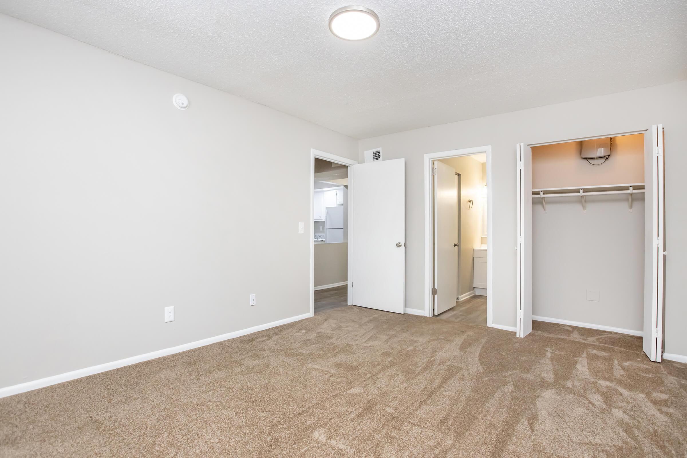A spacious, well-lit room with beige carpet and neutral-colored walls. The room features an open layout, a closet with sliding doors, and a doorway leading to a bathroom. Natural light is provided by a ceiling light fixture, creating a welcoming atmosphere.