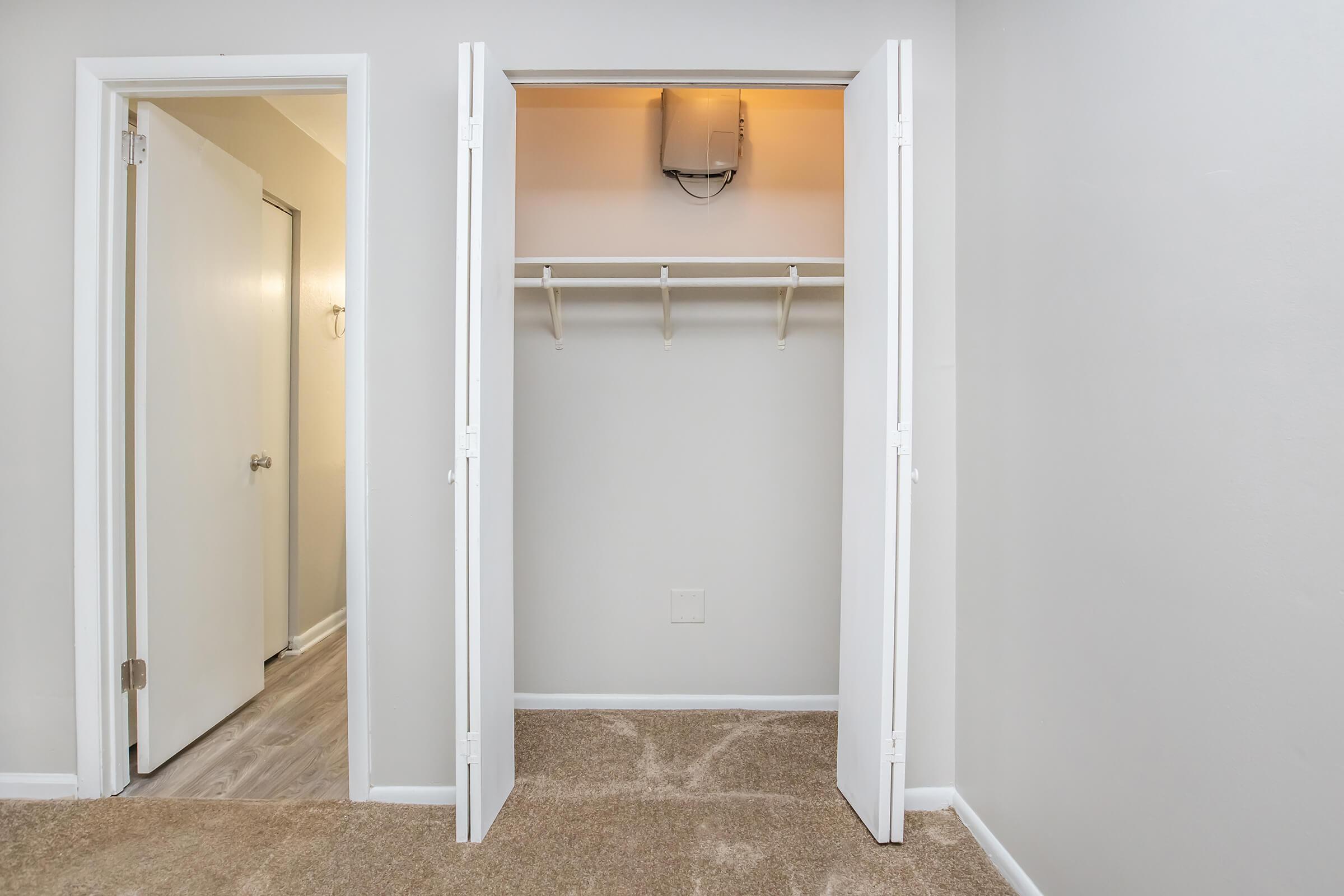 A well-lit, empty closet with two white doors slightly open, revealing a hanging rod and a small light fixture above. The walls are painted light gray, and the carpeted area in front is beige. A white door is visible on the left, leading to another room.