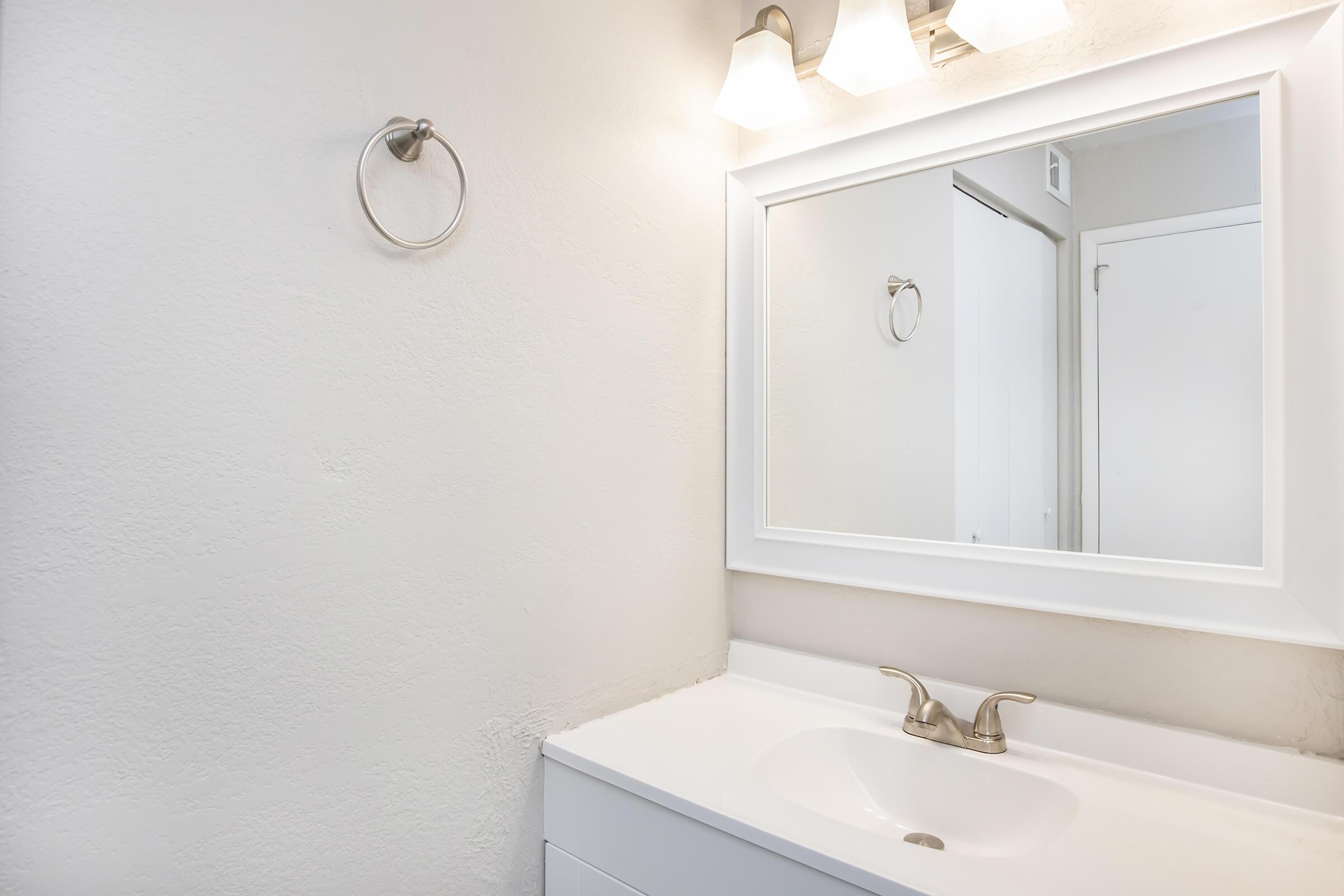 A clean, modern bathroom featuring a white sink and faucet, a rectangular mirror with a white frame, and a wall light fixture. The walls are painted in a light color, and there's a towel ring mounted on the wall. A door can be seen in the background, adding to the minimalist aesthetic.