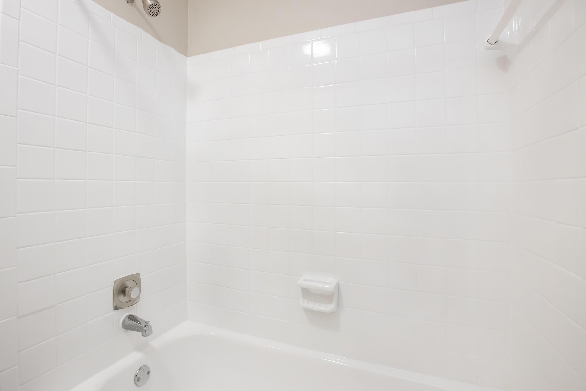 A clean, modern bathtub with white tiled walls. The bathtub has a chrome faucet and a hand shower attachment. There is a soap dish mounted on the wall, and the overall appearance is bright and minimalistic, giving a sense of cleanliness and simplicity.
