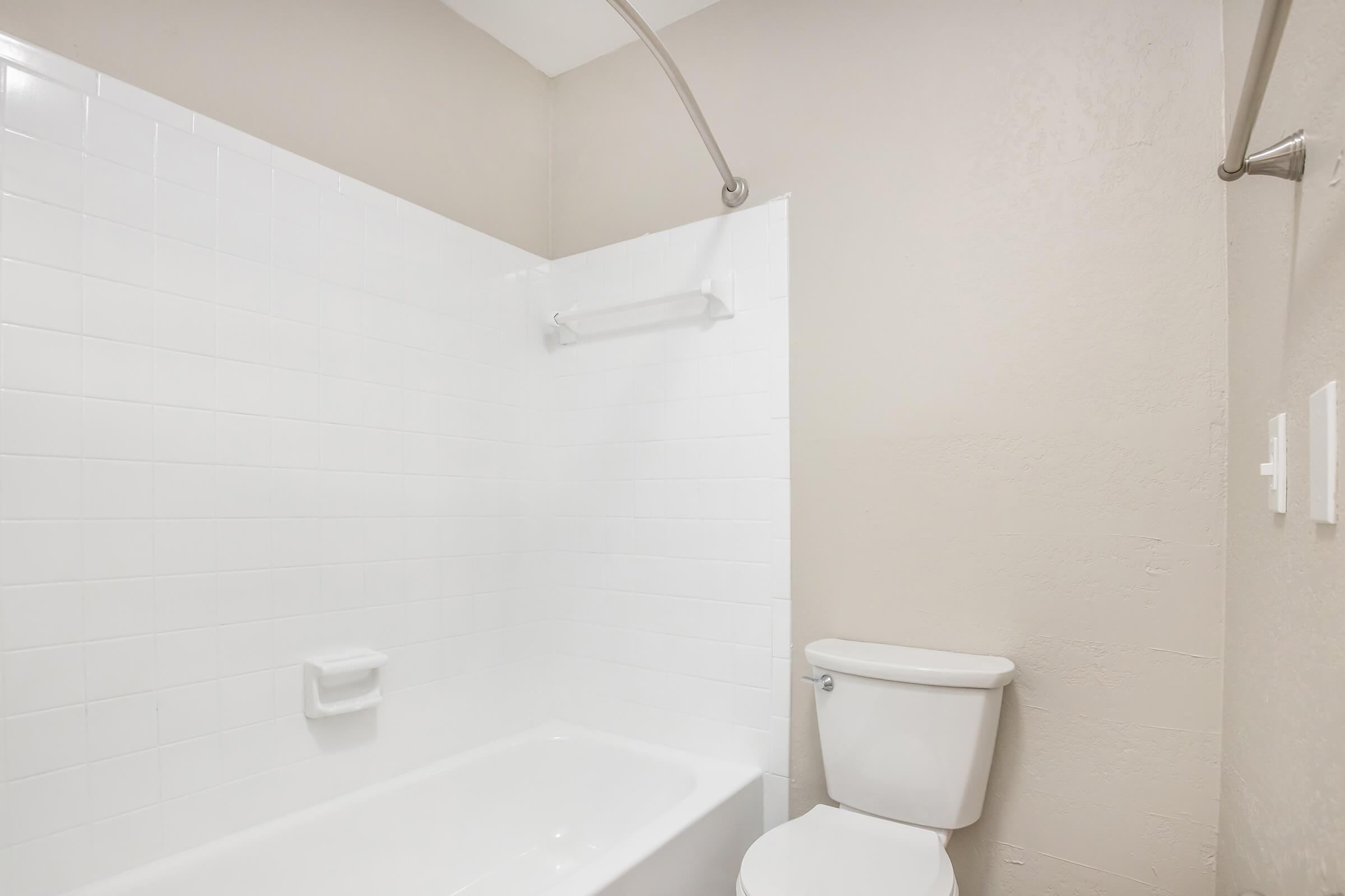 A clean bathroom featuring a white tiled shower area with a bathtub, a toilet beside it, and beige walls. The shower rod is visible, and there are grab bars installed. The overall design is minimalist and modern.