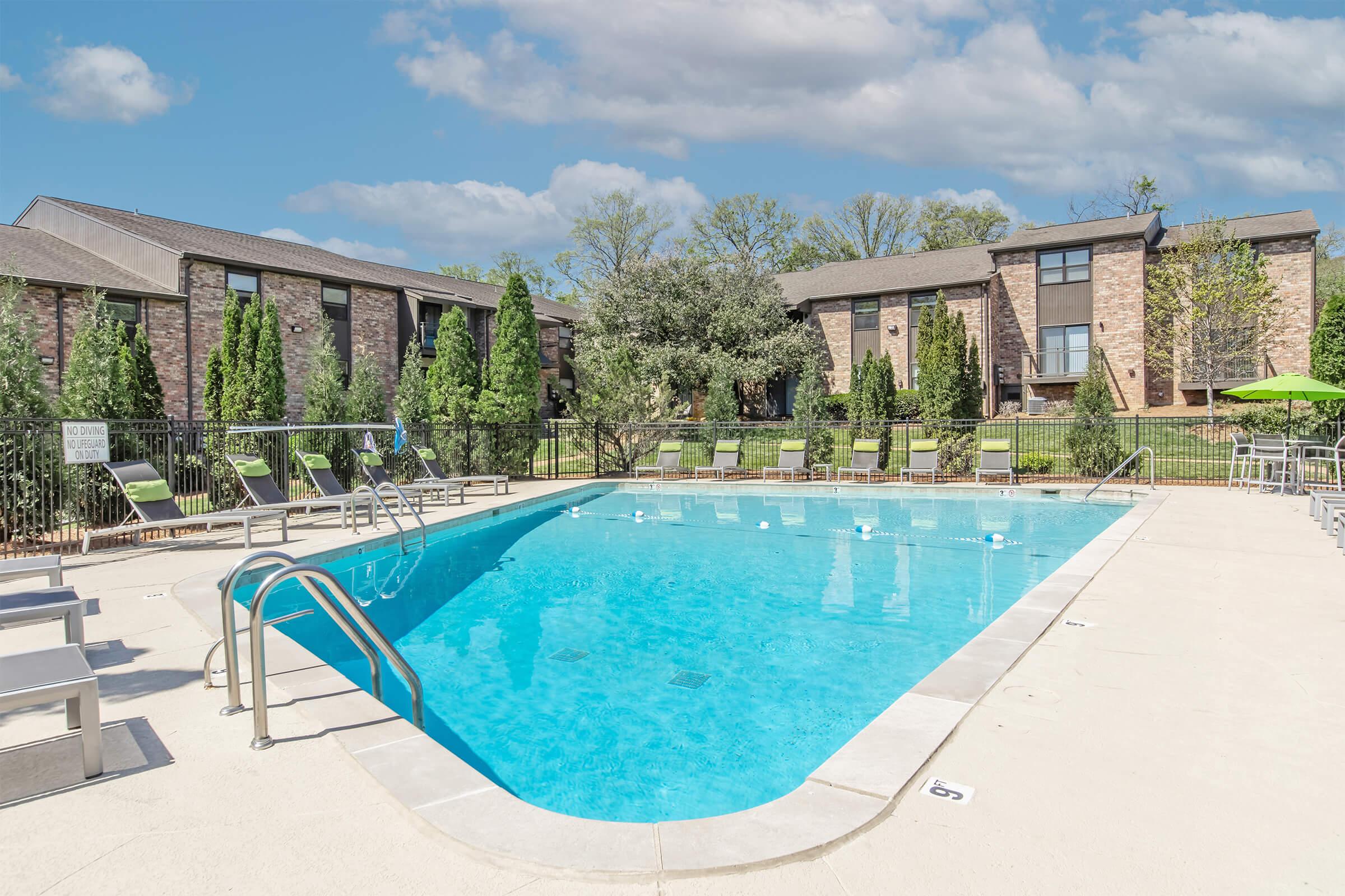 A clear blue swimming pool surrounded by lounge chairs, with well-maintained landscaping and two apartment buildings in the background. Green trees and shrubs add to the inviting atmosphere, and a few tables with umbrellas are visible nearby. The sky is partly cloudy, enhancing the scene.