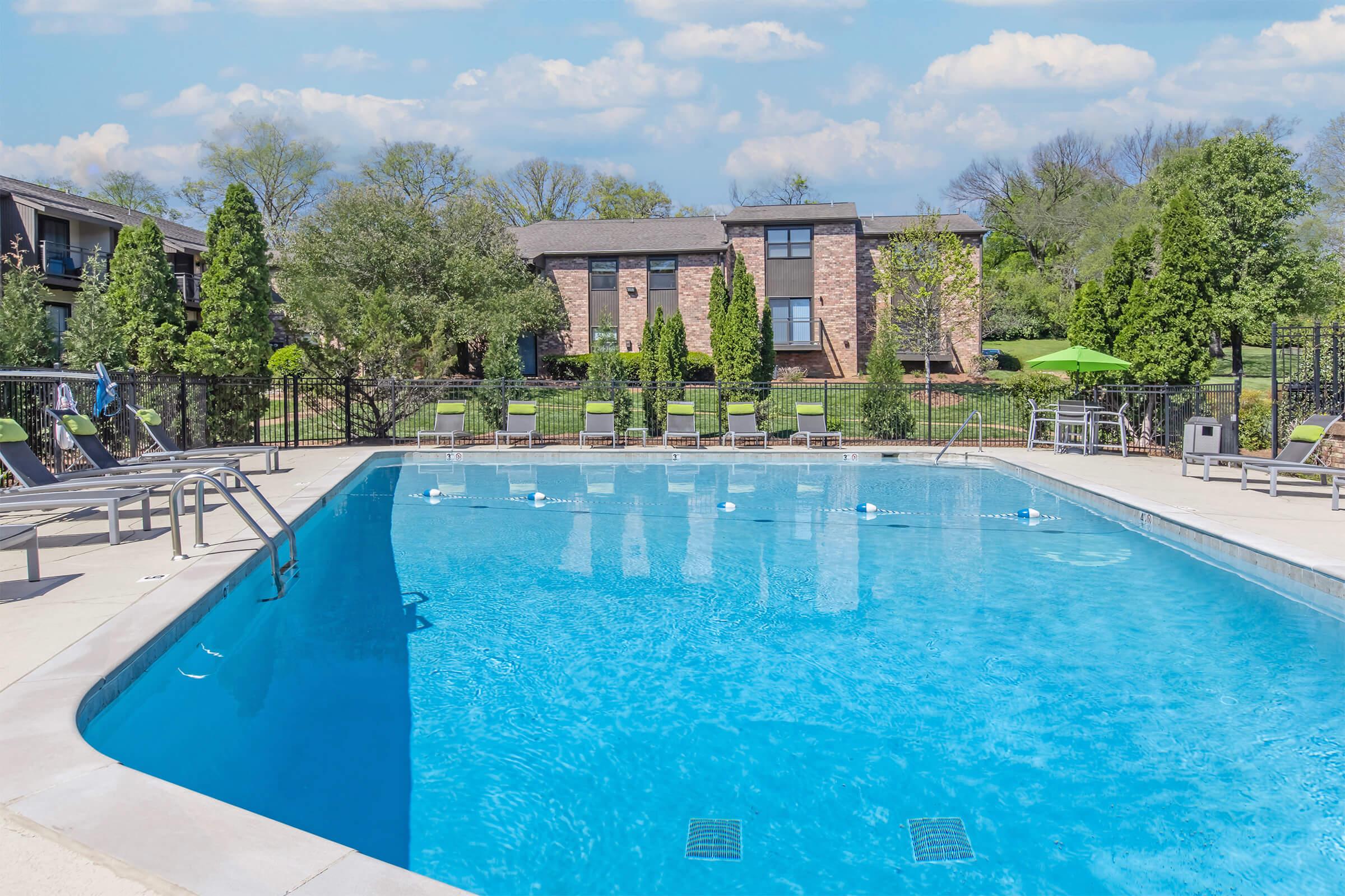 A clear blue swimming pool surrounded by lounge chairs, with lush green trees in the background. The pool area features a well-maintained deck and is adjacent to two brick apartment buildings, set against a bright sky with scattered clouds.