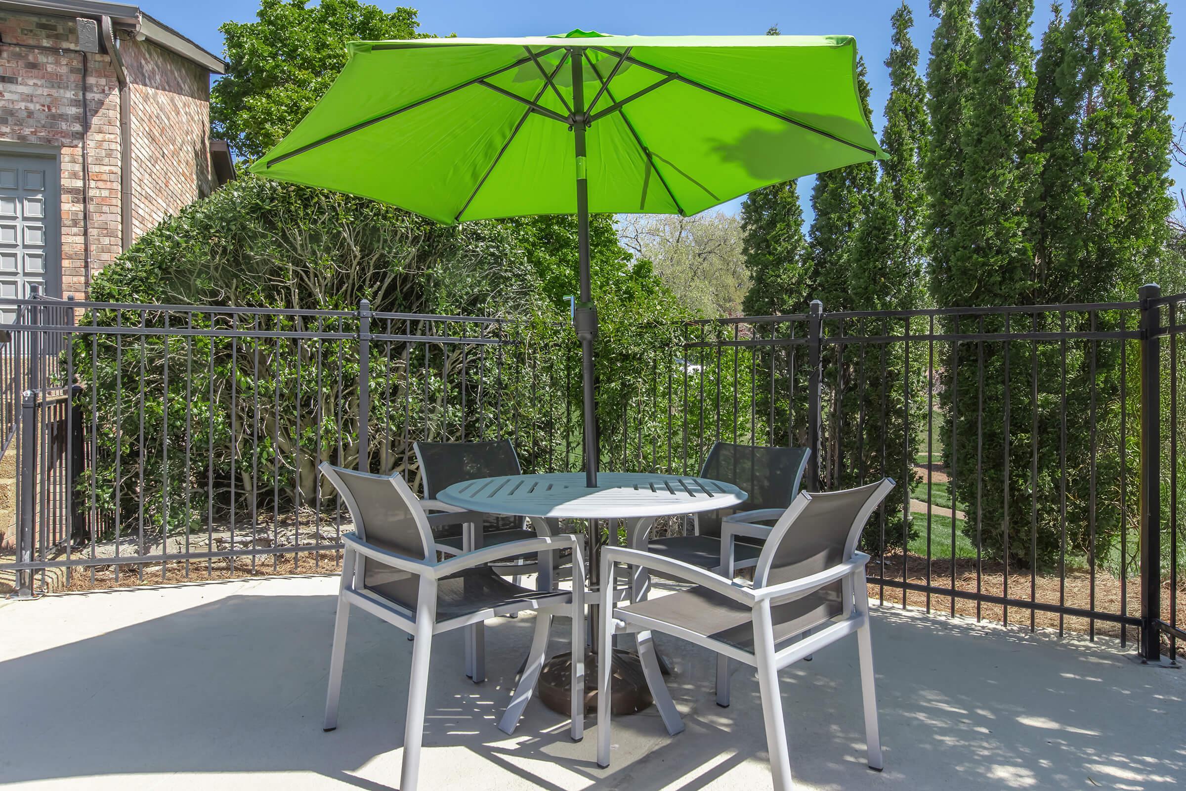 A patio area featuring a round table with four chairs under a bright green umbrella. The setting is surrounded by trees and a black iron fence, providing a cozy outdoor atmosphere. Sunlight casts a warm glow on the concrete surface.