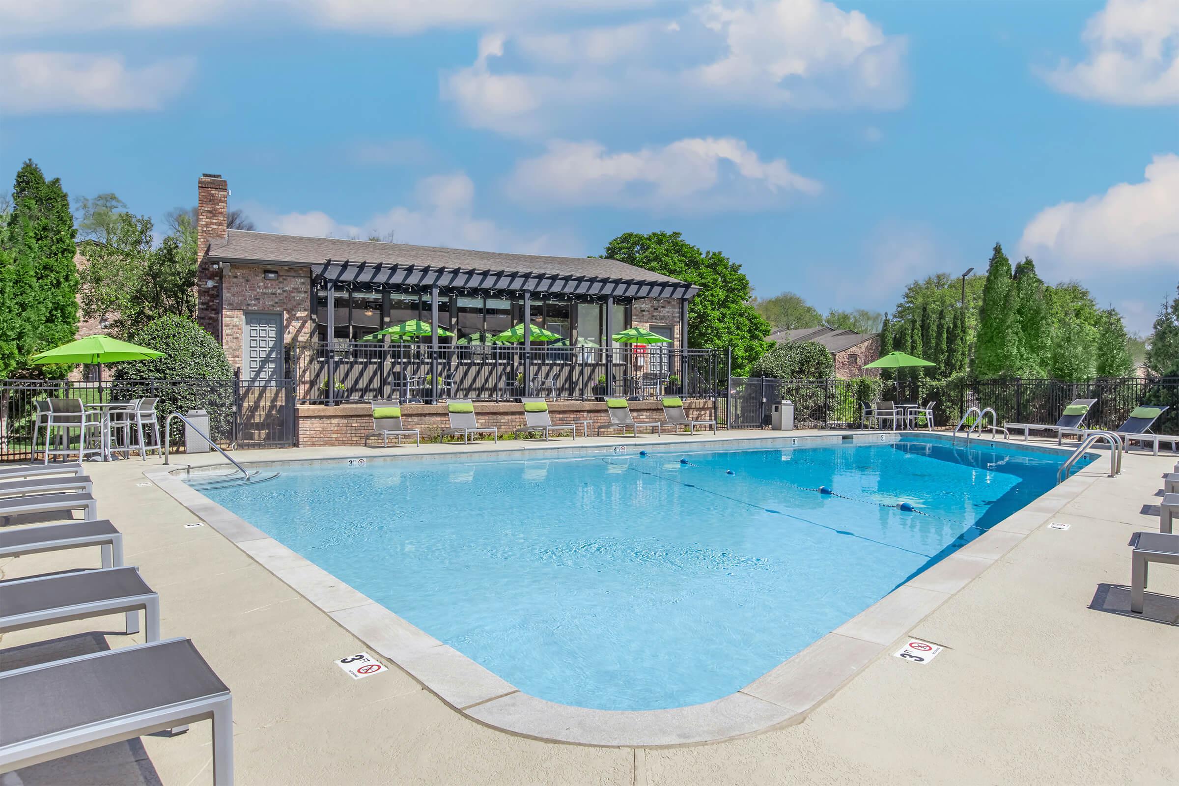 A sunny outdoor swimming pool surrounded by lounge chairs, with a building in the background featuring a shaded patio area and green umbrellas. Lush greenery frames the scene, creating a relaxing atmosphere ideal for leisure and recreation.