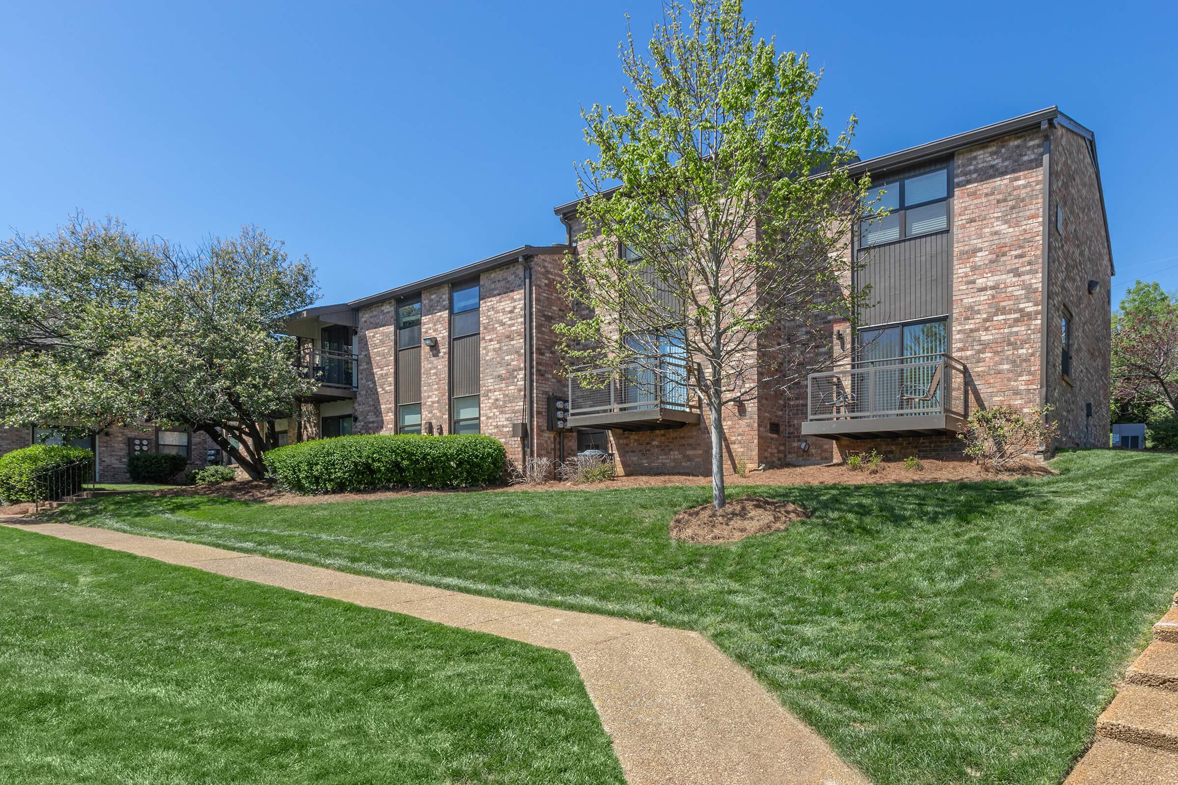A well-maintained brick apartment building surrounded by lush green grass and small trees. A pathway winds through the lawn leading to the entrance of the building, which features balconies on the upper floors. Clear blue sky adds to the inviting atmosphere.