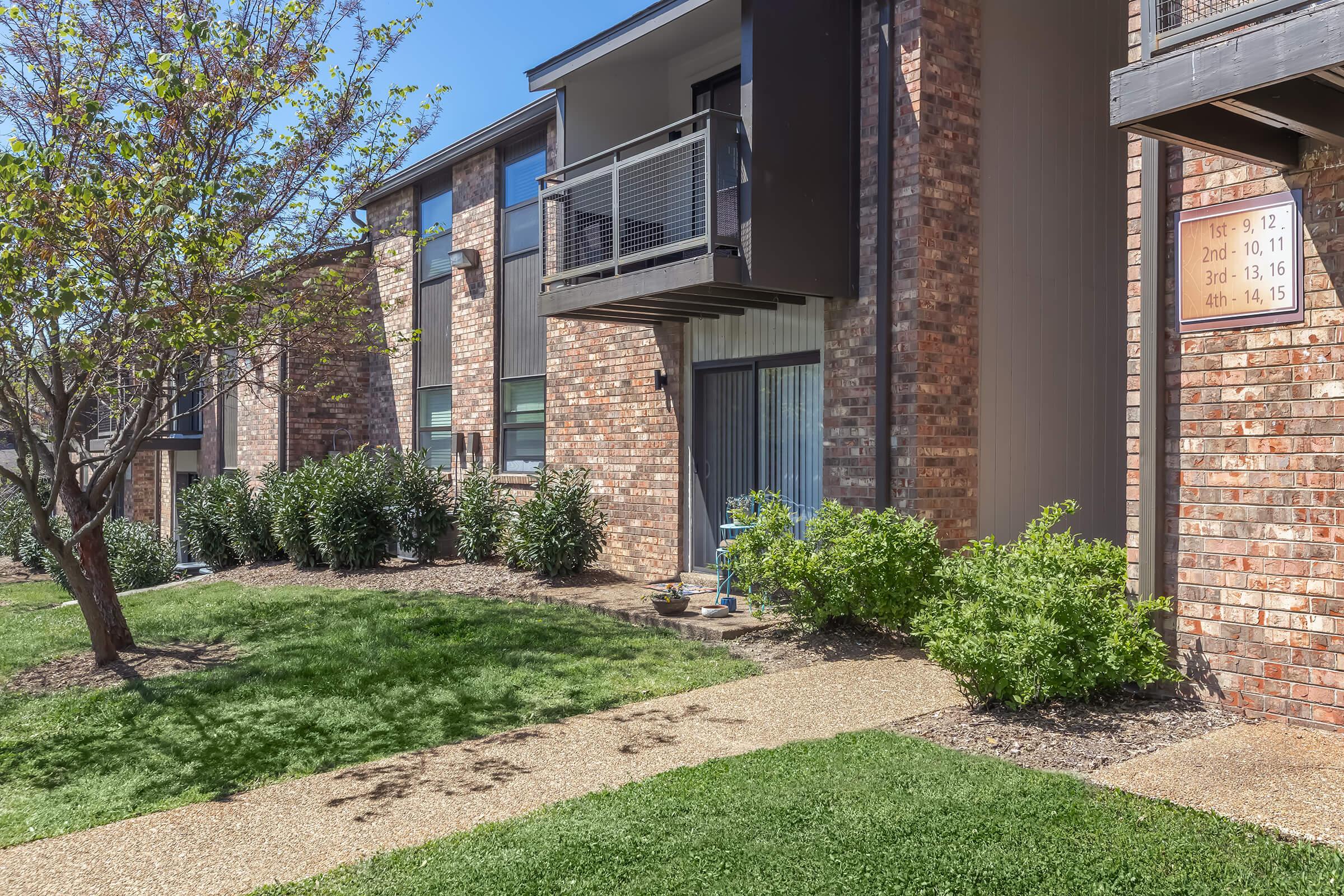 View of a residential apartment complex featuring brick facades, a small balcony, and well-maintained landscaping with shrubs and a tree. A pathway leads through the greenery, and a building directory is visible on one wall indicating apartment numbers. The sky is clear and sunny.