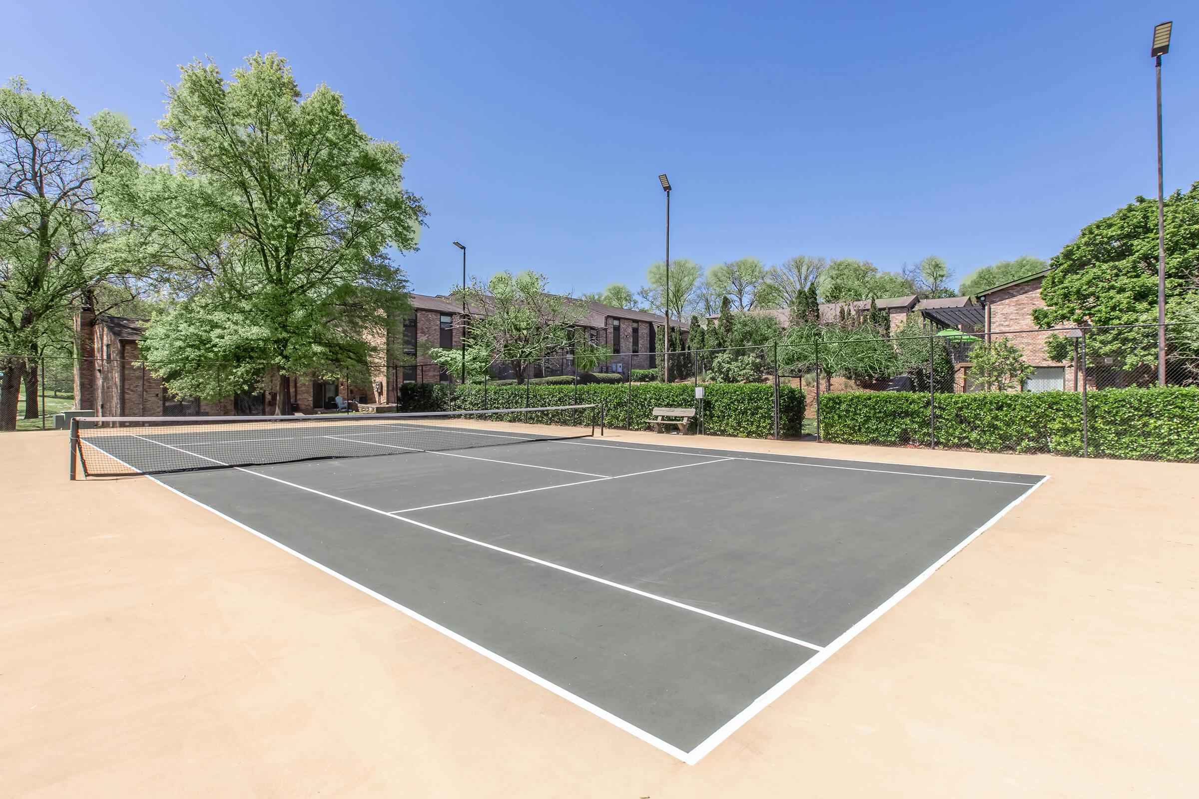 A well-maintained outdoor tennis court with a clay surface, featuring neatly marked lines and a net in the center. Surrounding the court are green trees and neatly trimmed hedges, with residential buildings visible in the background under a clear blue sky.