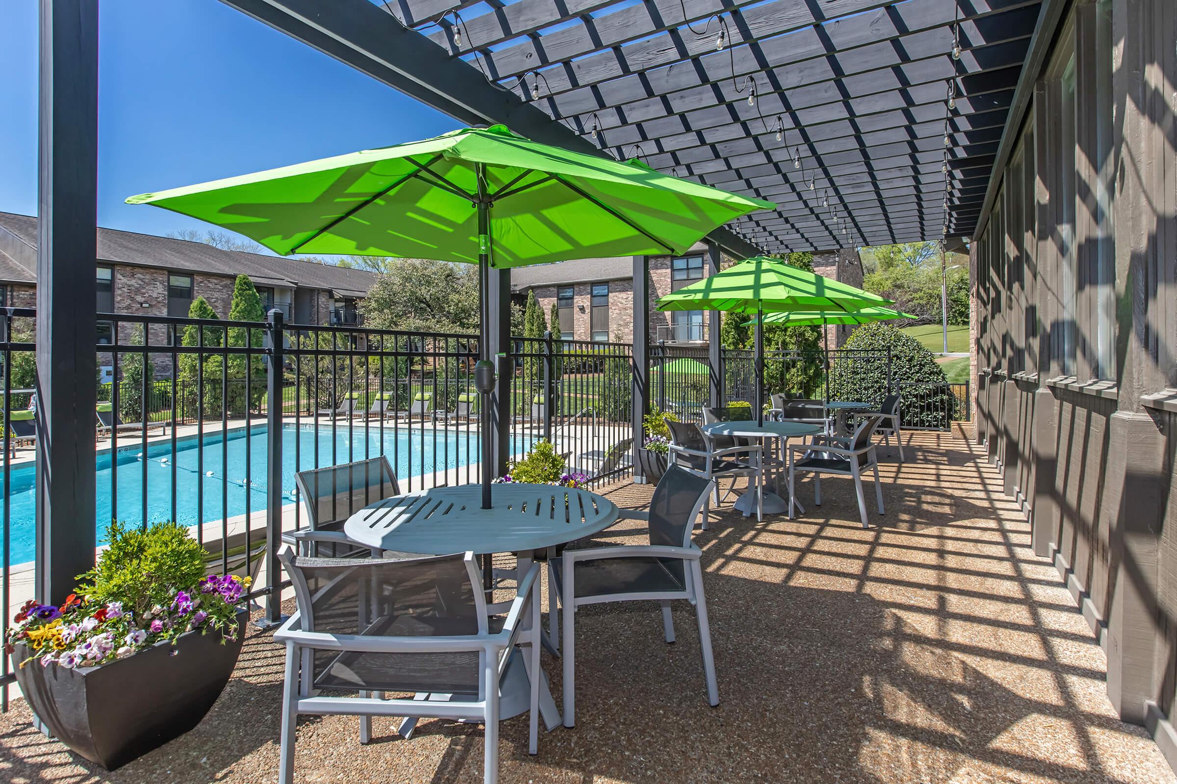 A sunny outdoor patio area featuring several tables and chairs under vibrant green umbrellas. Potted plants add greenery to the space, which overlooks a clear blue pool surrounded by lush landscaping. The setting is ideal for relaxation and dining.